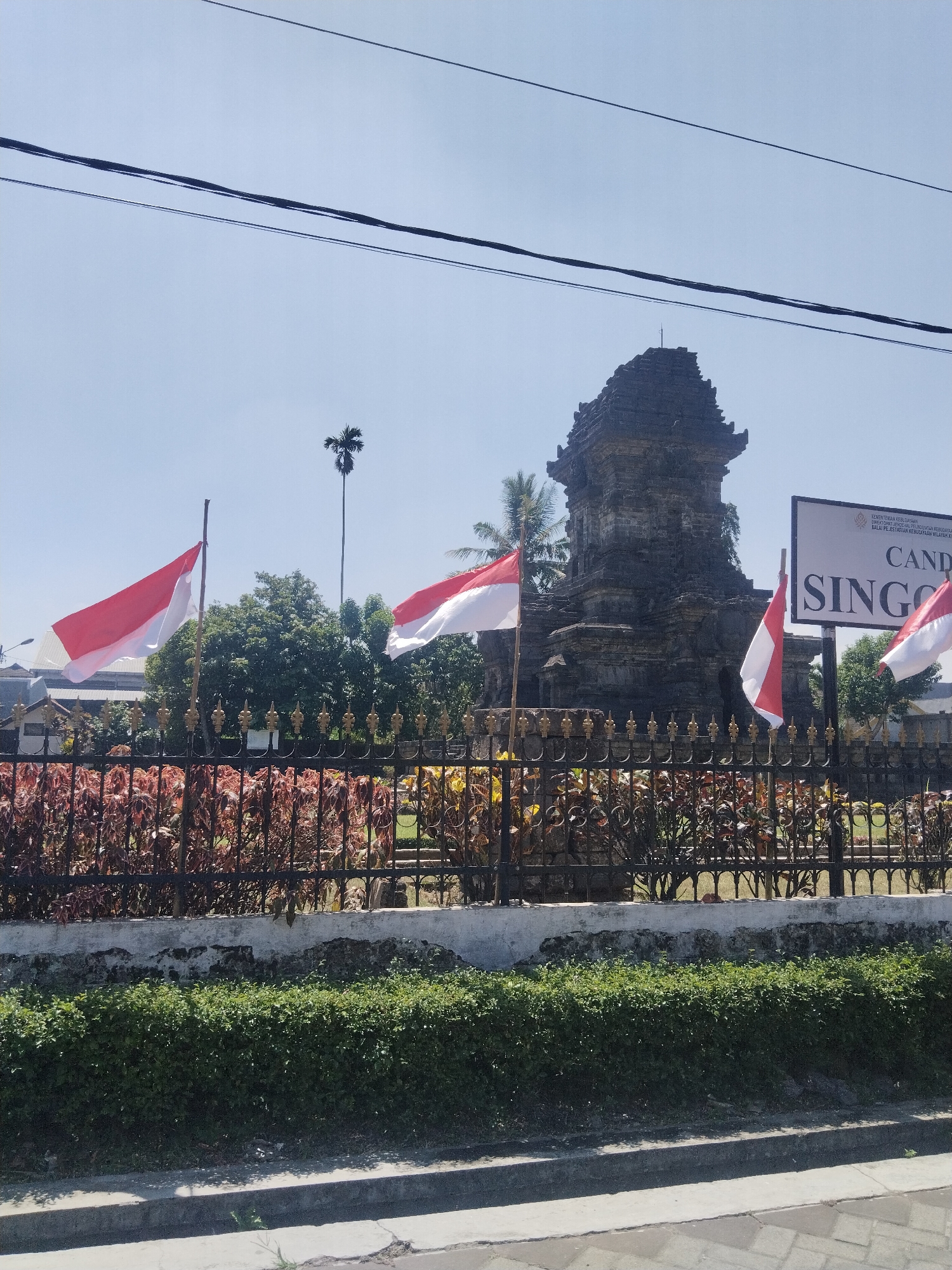 A historic stone structure, possibly a temple or monument, stands under a clear blue sky in Candirenggo, Indonesia. Several red and white Indonesian flags are displayed prominently on flagpoles in front of the structure. A black metal fence with ornate gold accents encloses the monument, behind which is a garden with various plants. A sign with Indonesian text, including "CANDI SINGO" is visible to the right of the monument. Overhead power lines crisscross the sky above the scene. The area in front of the fence appears to be a street or walkway, with a concrete curb and road surface.