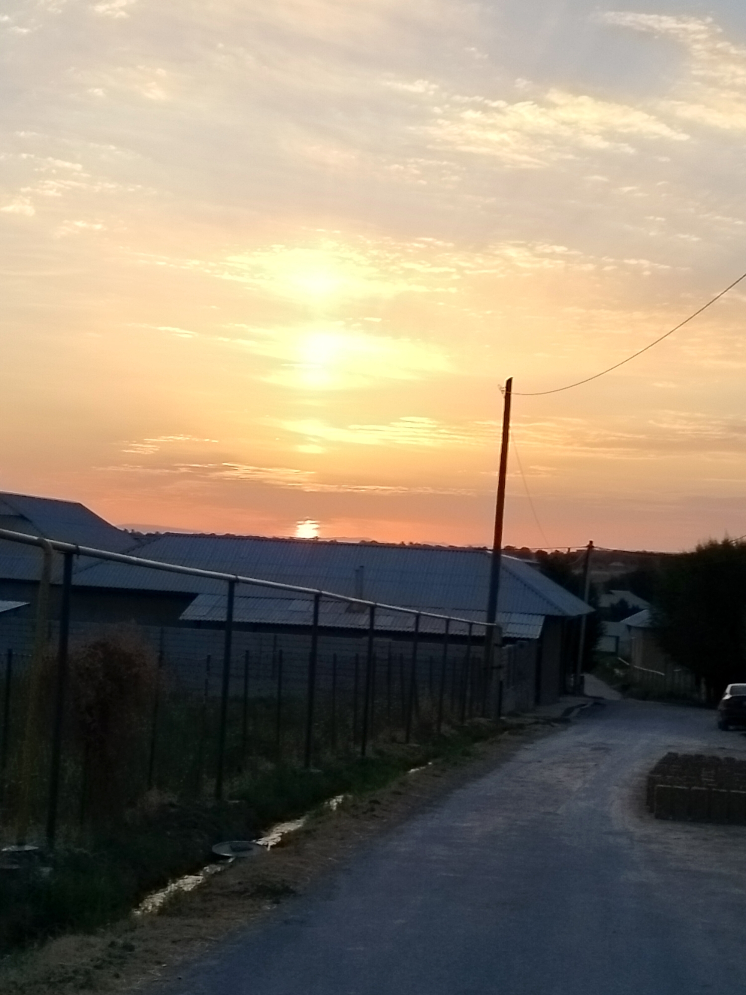 An outdoor, vertical, full shot depicts a road leading into a rural settlement at sunset. The sky is filled with wispy clouds and illuminated by a bright, orange sun positioned near the horizon. In the foreground, a dirt road curves to the right. To the left of the road, a wire fence runs along a strip of dry grass and a small ditch. Beyond the fence, buildings with corrugated metal roofs are visible. A tall wooden pole stands in the middle ground, with a wire extending from its top. In the background, more buildings and trees are scattered throughout the landscape, shrouded in the fading light. The overall atmosphere is tranquil and serene, with the warm colors of the sunset creating a peaceful ambiance.