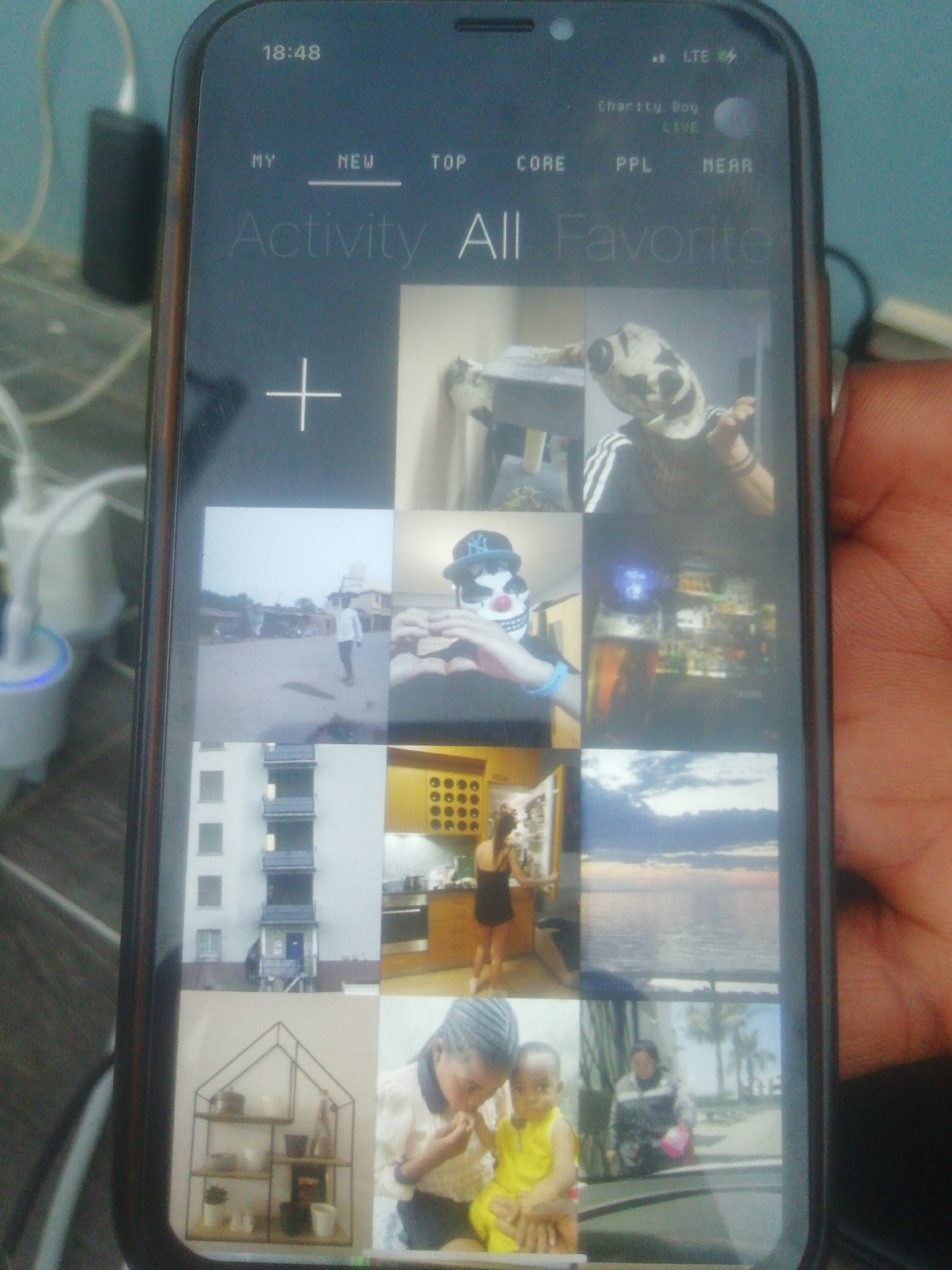 A phone screen displays a collage of images under the headings "My, New, Top, Core, PPL, Near, Activity All Favorite." The phone's time reads 18:48, and it's connected to LTE. The collage includes images of: a person in a mask; a cityscape; the interior of a building; an ocean scene; a woman in a kitchen; a shelf of decorative items; and a child being held by an adult. Based on these images and the reference to 'Charity Boy', the location may be Ikorodu, Nigeria. Other objects visible are phone chargers and cables.