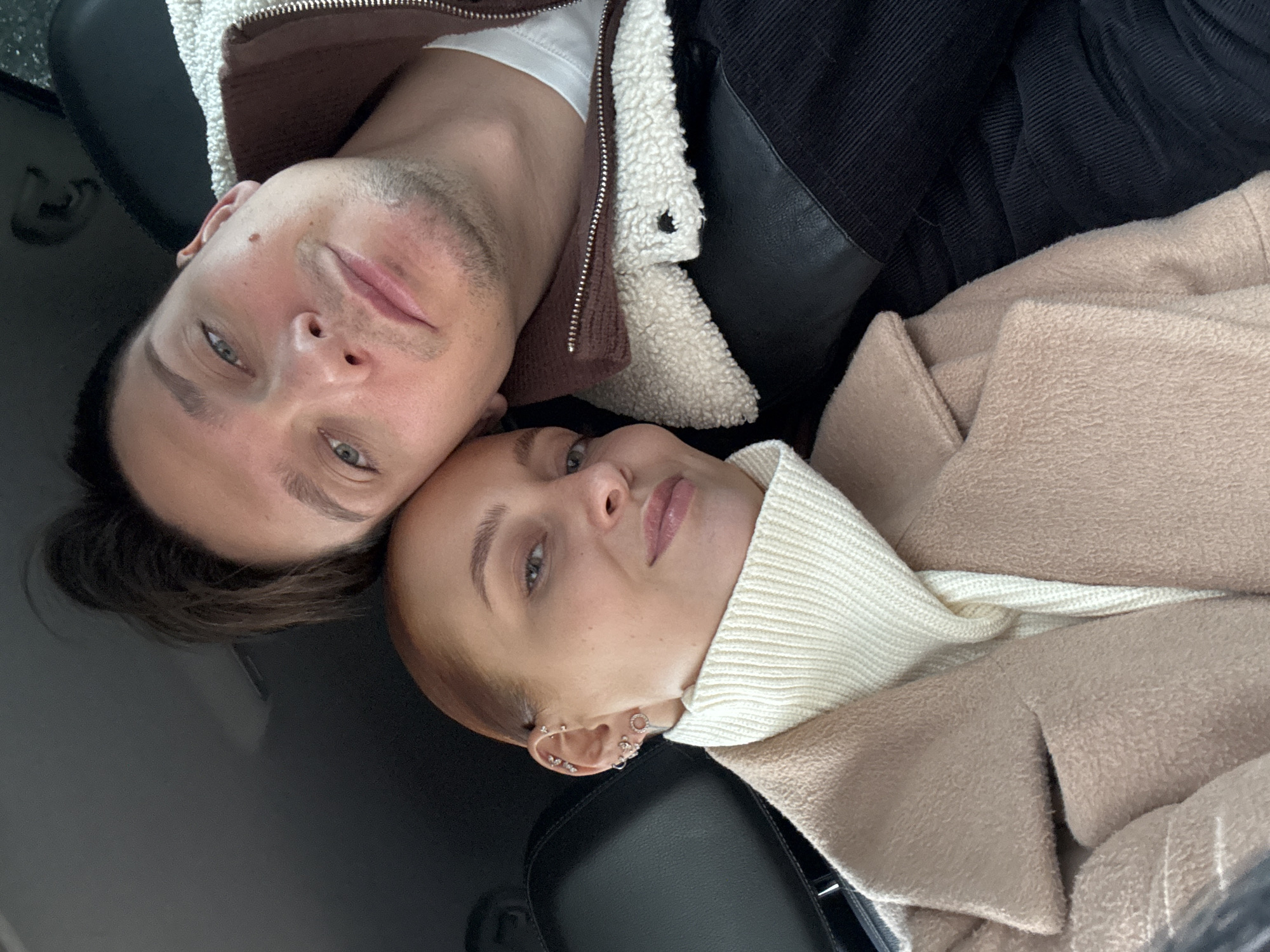 The image is a close-up, overhead shot of two people lying down. The person on the left is male, with short brown hair, light eyes, and fair skin. He is wearing a white t-shirt under a brown jacket with a white, sherpa-lined collar. The person on the right is female, with red hair, fair skin, and multiple piercings in her left ear. She is wearing a cream-colored turtleneck sweater and a tan coat. The background is dark and appears to be the interior of a vehicle. There is no information available that specifies the location of Chaikovsky, Russia in the image.