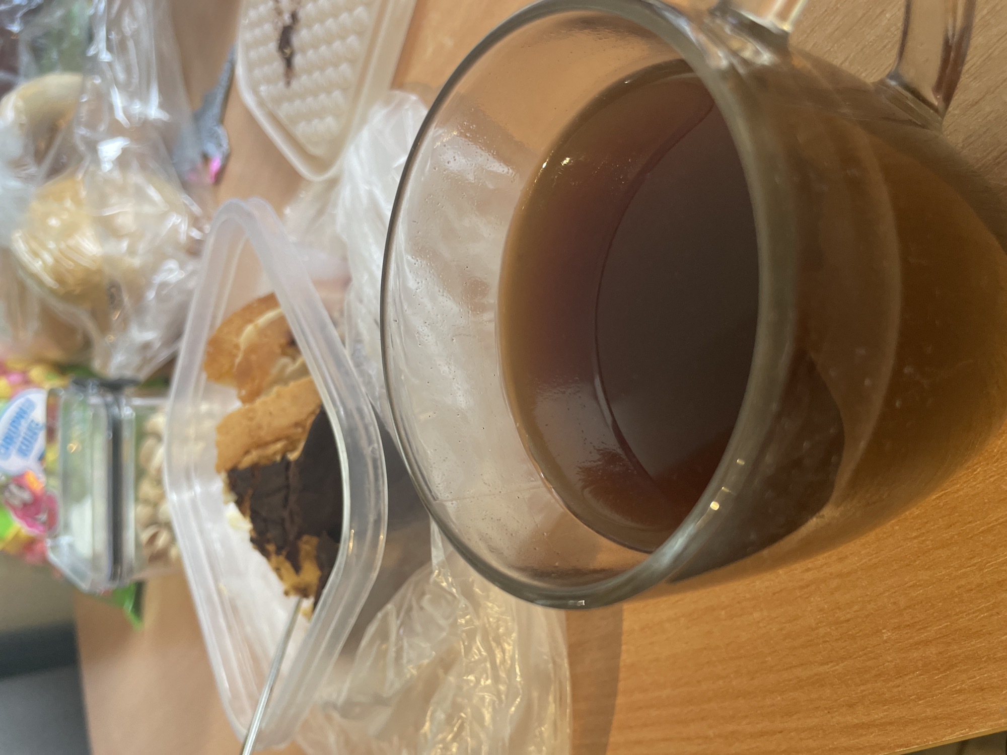 A close-up, eye-level shot shows a clear glass mug filled with dark brown liquid, likely coffee or tea, resting on a light brown wooden table. The mug has a visible handle. In the background, partially obscured, are several food items in clear plastic containers and bags, including what appears to be a slice of cake with chocolate frosting, some cookies or pastries, and a container of nuts. The environment appears to be indoors, possibly a home or office.