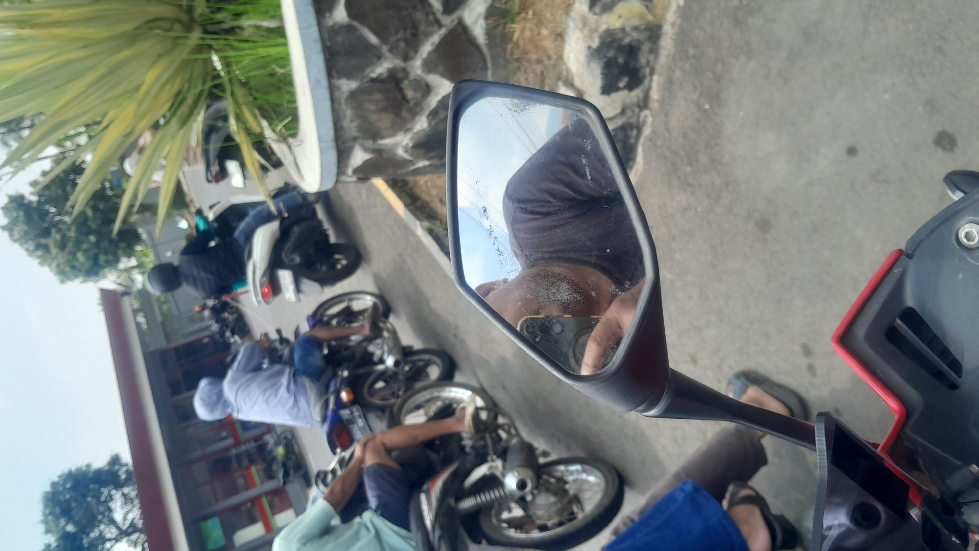 The image depicts a street scene in Subang, Indonesia, likely near a commercial establishment. Several motorcycles are visible, with a man in a light blue hooded jacket and shorts riding one in the foreground. Behind him, another person wearing a black jacket and helmet is on a white scooter. To the left, a person in a light blue shirt and shorts is seated on a motorcycle. The perspective suggests the photo is taken from the point of view of someone on a motorcycle, as a motorcycle mirror is prominently featured, reflecting the face and upper body of the rider, who is wearing a dark t-shirt and has a beard. In the background, a building with a reddish-brown roof and glass windows appears to be a shop or service station. Lush green vegetation and a stone-clad wall are also visible on the right side of the frame. The road surface is asphalt, and the sky is overcast.