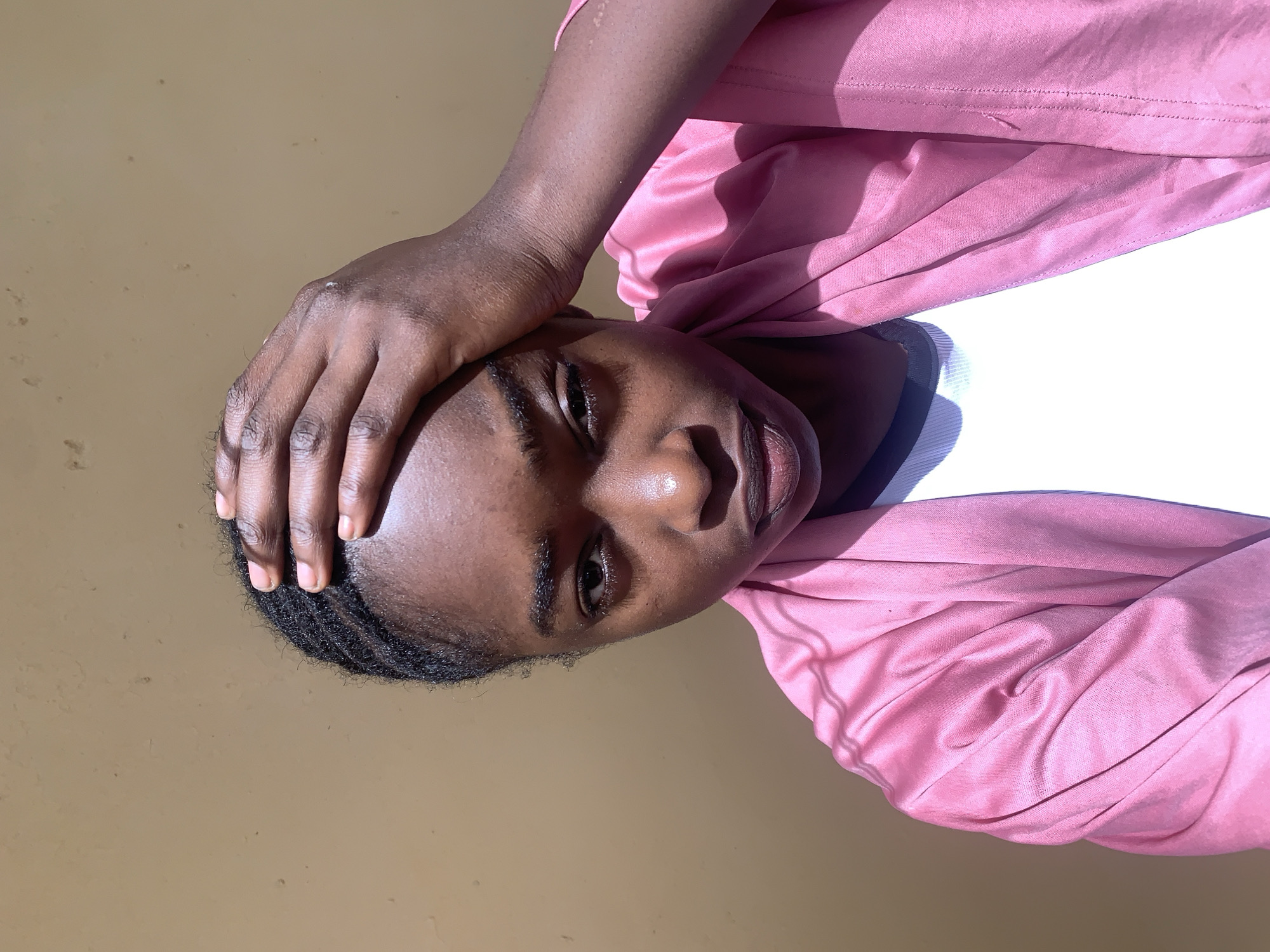 A young person with dark skin and braided hair is the primary subject of the photograph. They have their right hand resting on their head. The individual is wearing a white top underneath a pink jacket or cardigan. The background is a solid, pale brown color. The image appears to be taken indoors under sunlight. There is no visible evidence that indicates Rafin Par, Nigeria.