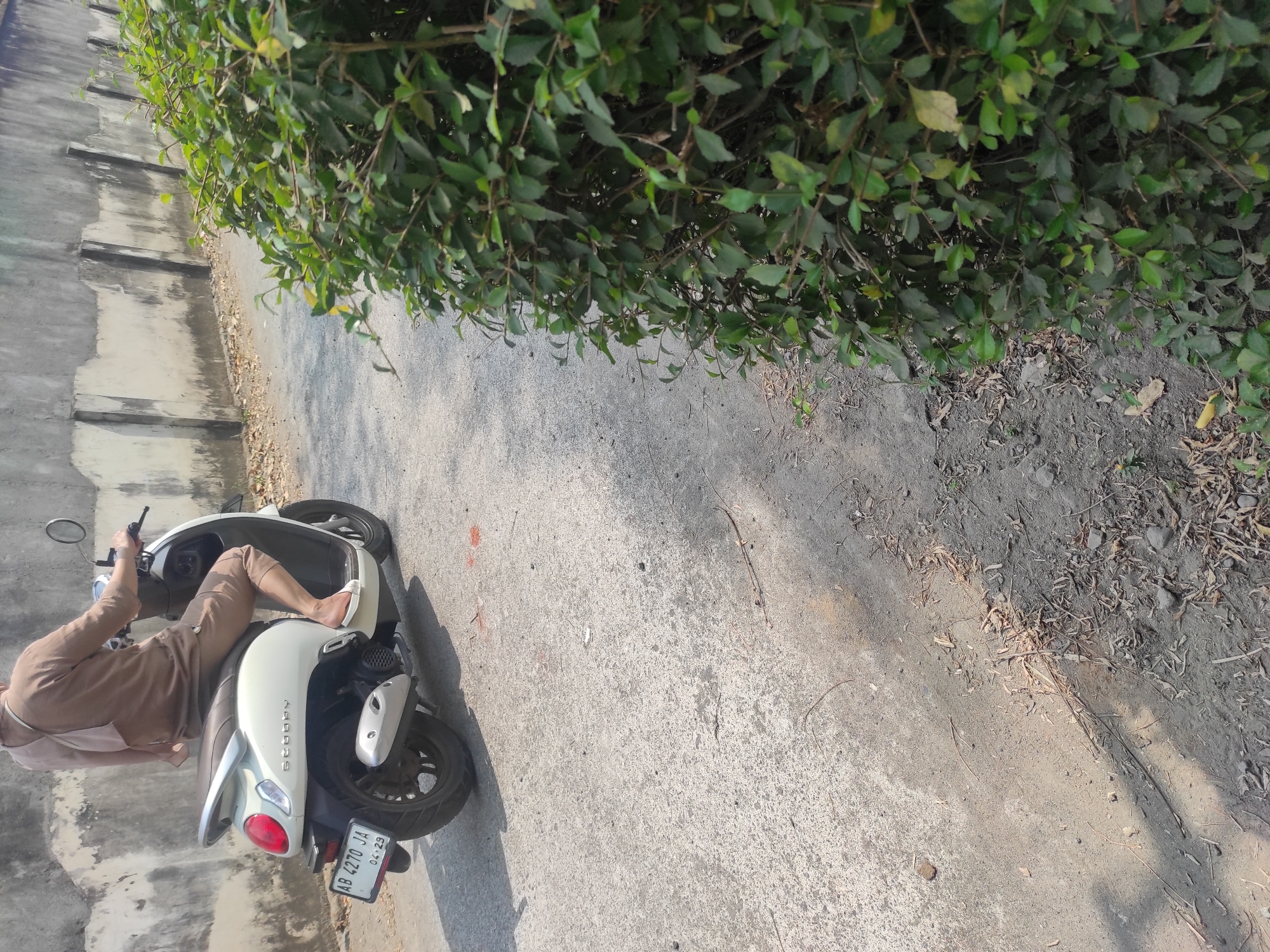 A person wearing a brown hijab and brown clothing rides a white scooter with a license plate reading "AB 4270 JA" in Pleret, Indonesia. The scooter has "Scoopy" written on its side. The scooter is on a paved road next to a grey concrete wall and a dense green bush. The ground is covered in dirt, leaves, and small stones. The scene is illuminated by sunlight, casting shadows on the road.