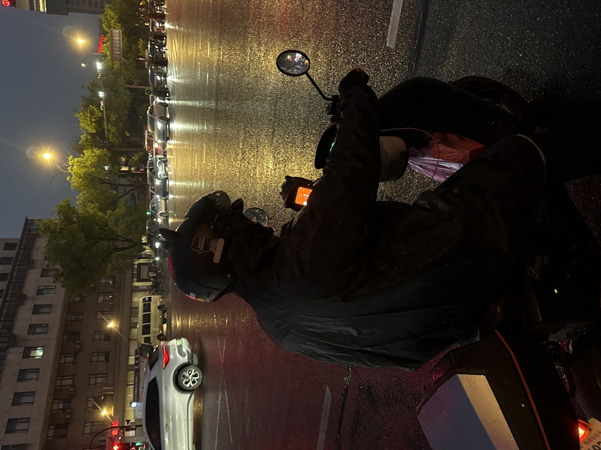The image depicts a rainy evening scene on a city street in Yiwu, China. A person wearing a black helmet and a black raincoat is seated on a scooter, facing away from the camera. The scooter has a purple plastic bag hanging from it. The street is wet and reflects the surrounding lights, indicating it has been raining. In the background, several vehicles, including cars and a white bus, are visible on the road, some with their headlights on. Buildings line the street, with lights on in some of the windows. The overall atmosphere is one of a typical urban environment during inclement weather.