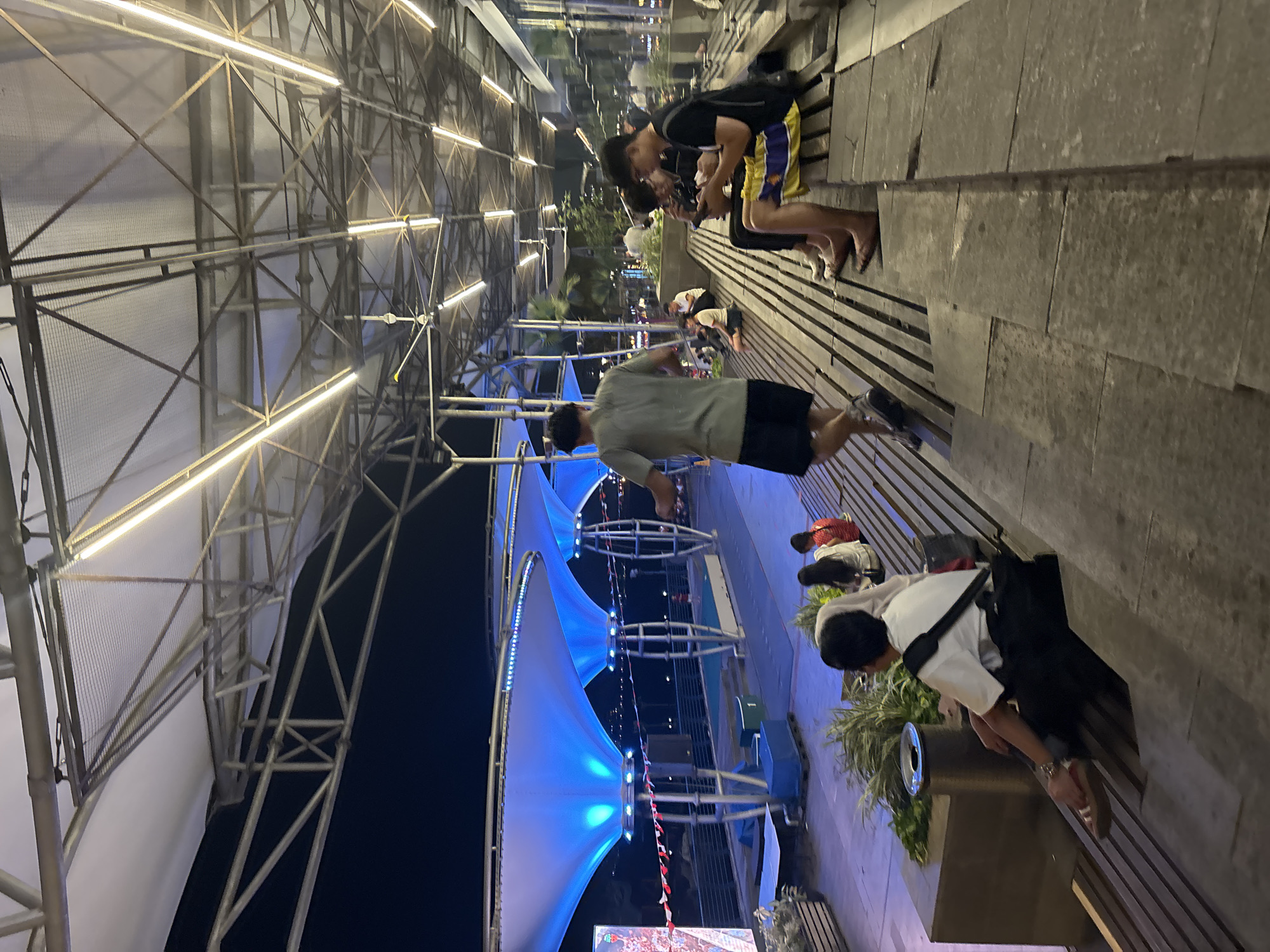 The image depicts an outdoor seating area at night, likely a public space in North Jakarta, Indonesia, possibly Ancol. The scene is illuminated by overhead artificial lights and the blue glow from large, tent-like structures. In the foreground, several people are seated on tiered wooden benches. To the left, a person in a white shirt and black pants is looking down. Further to the right, two young men are engrossed in their mobile phones, with one wearing a black t-shirt and shorts with a colorful design, and the other in a black t-shirt and pants. Behind them, more individuals are seated, some appearing to be in conversation. A man in a grey t-shirt and black shorts is walking away from the camera. The background reveals a waterfront area with lights and structures, suggesting a developed urban environment.