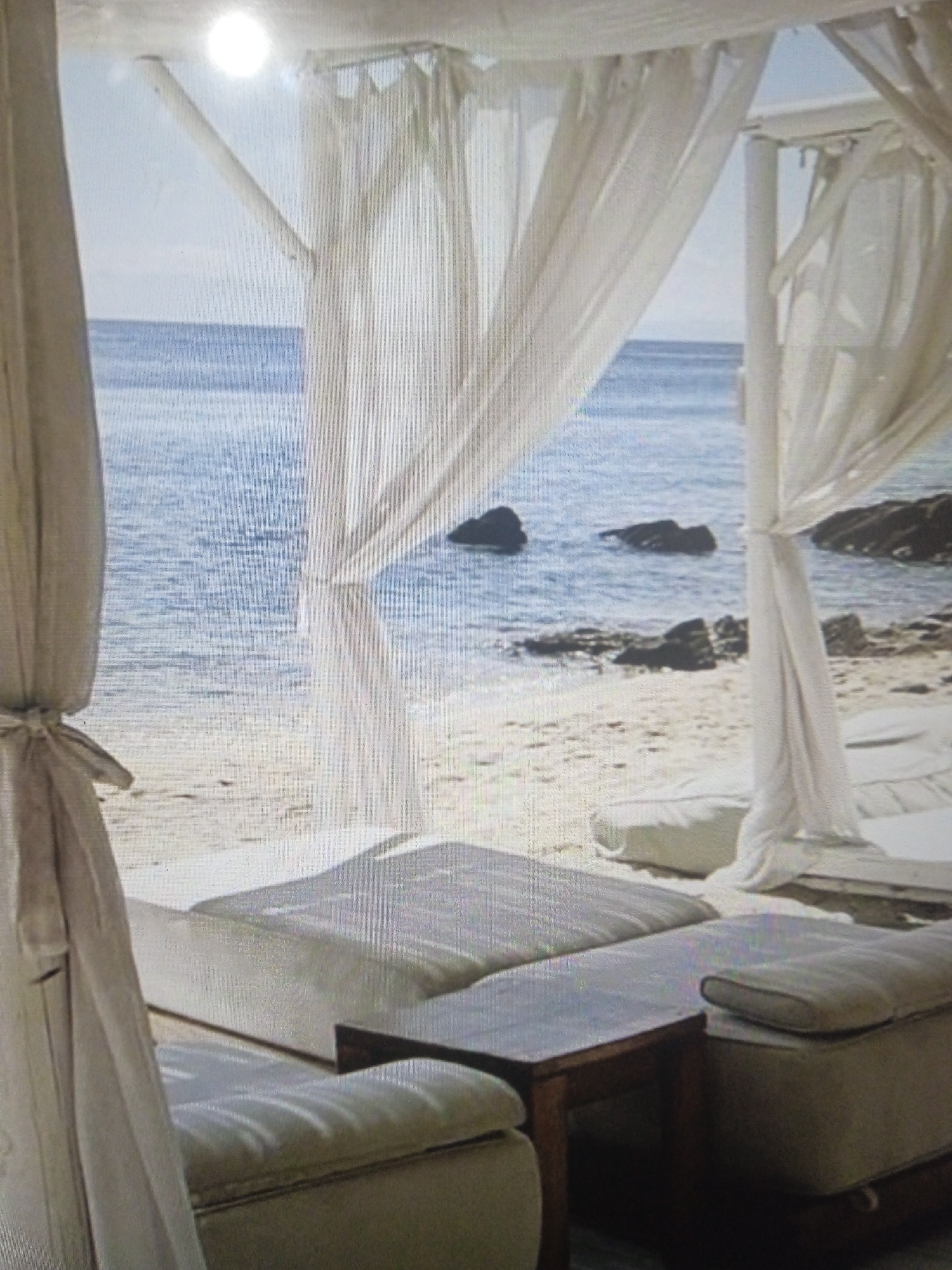 The image shows an outdoor seating area on a sandy beach, with a view of the ocean and rocks. The seating area is covered by white sheer curtains, which are draped from a structure above. In the foreground, there are several white cushioned seating platforms and a small wooden table. The ocean is blue and calm, with a few rocks visible in the water. The sand is light-colored. There are no people visible in the image. The scene appears to be a tranquil resort or private beach area. It is not possible to determine the location of this scene within Chernivtsi, Ukraine, as Chernivtsi is an inland city and does not have a coastline.