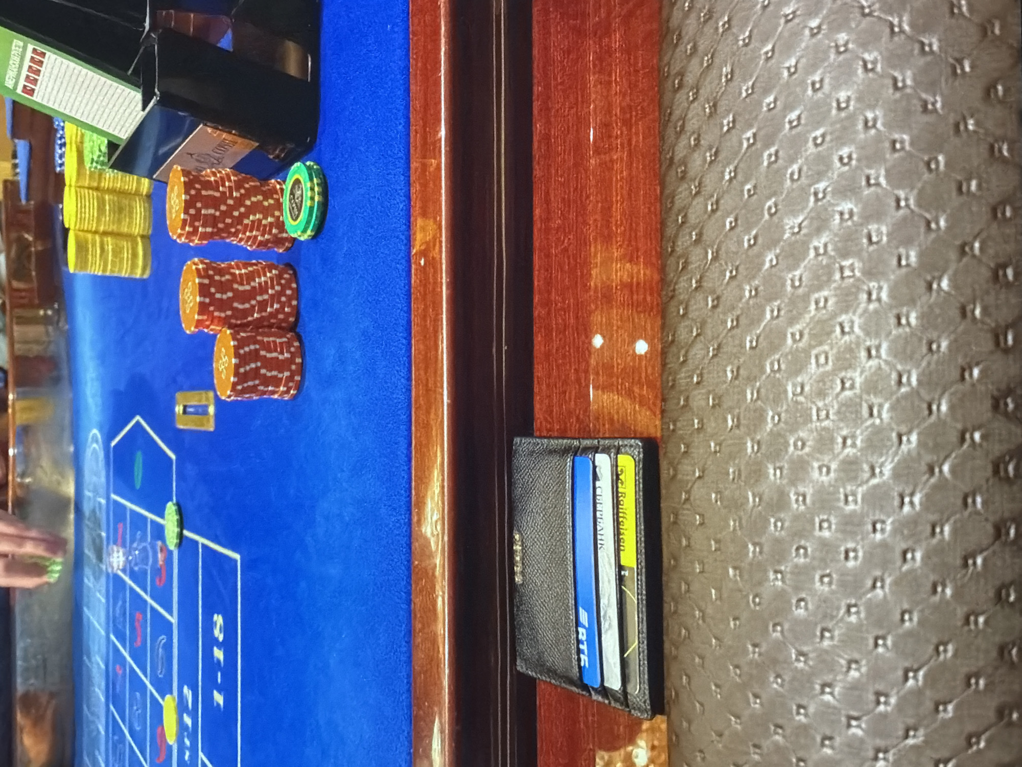 This image shows a roulette table in an unknown city. The table is covered in blue felt with roulette betting lines. Several stacks of casino chips are placed near the edge of the table. A cardholder is positioned at the edge of the table. The table is bordered by wood trim and a patterned, possibly upholstered surface. A hand is visible on the left side of the image.