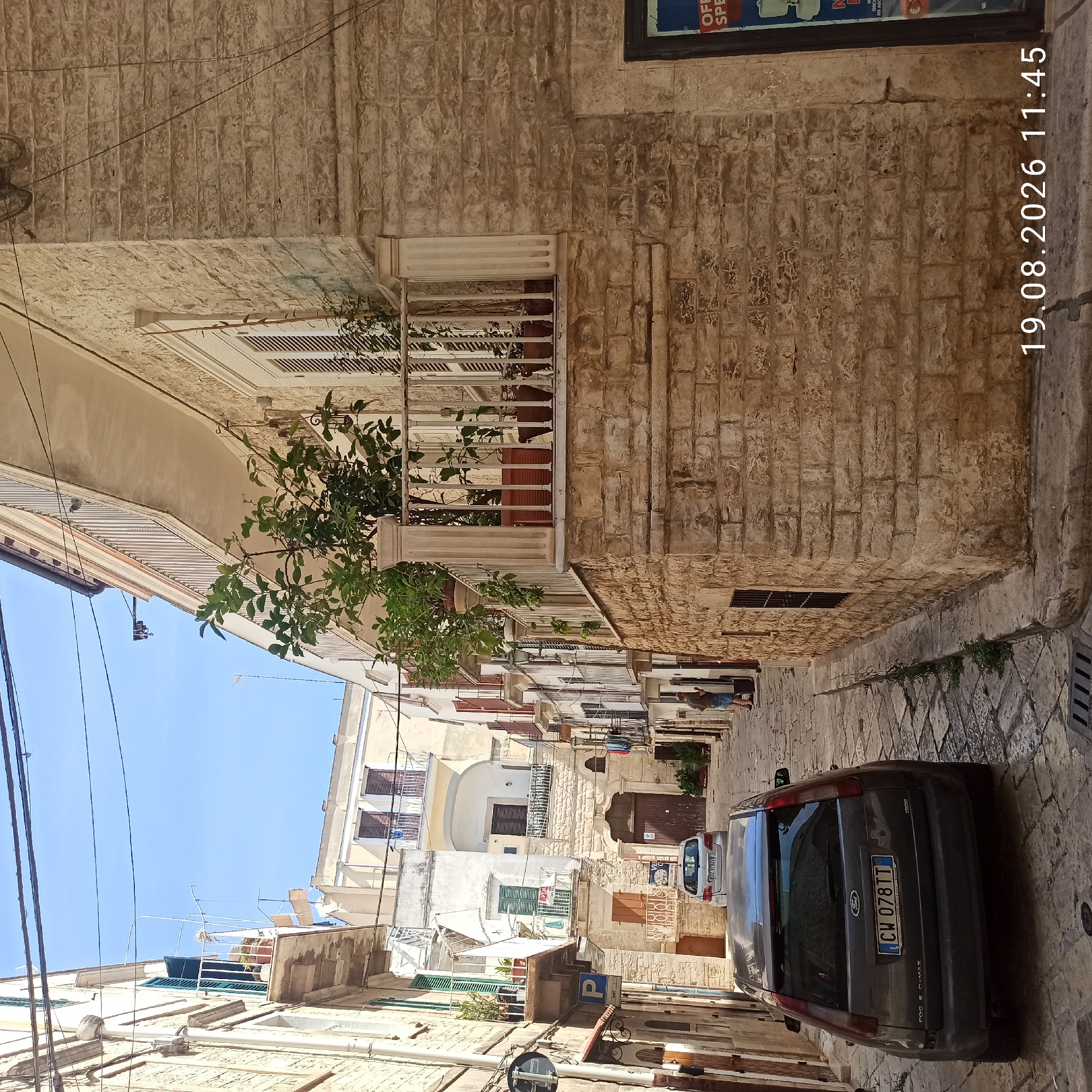 This is a forensic, matter-of-fact description of the image:
The image depicts a narrow street in Ruvo di Puglia, Italy, characterized by aged stone buildings and cobblestone pavement. In the foreground, a dark gray Ford C-Max hatchback is parked on the left side of the street, facing away from the viewer. Its license plate is visible, reading "CW 078TT". Further down the street, another silver car is parked. The buildings lining the street are constructed from light-colored stone blocks, with multiple levels featuring balconies and windows. Some balconies are adorned with potted plants. A man in a green shirt and blue shorts stands on the right side of the street, facing a doorway. Electrical wires are strung across the upper portion of the scene. A street sign with a blue 'P' symbol is visible on the left. The timestamp "19.08.2026 11:45" is overlaid on the bottom right of the image. The sky is clear and blue.