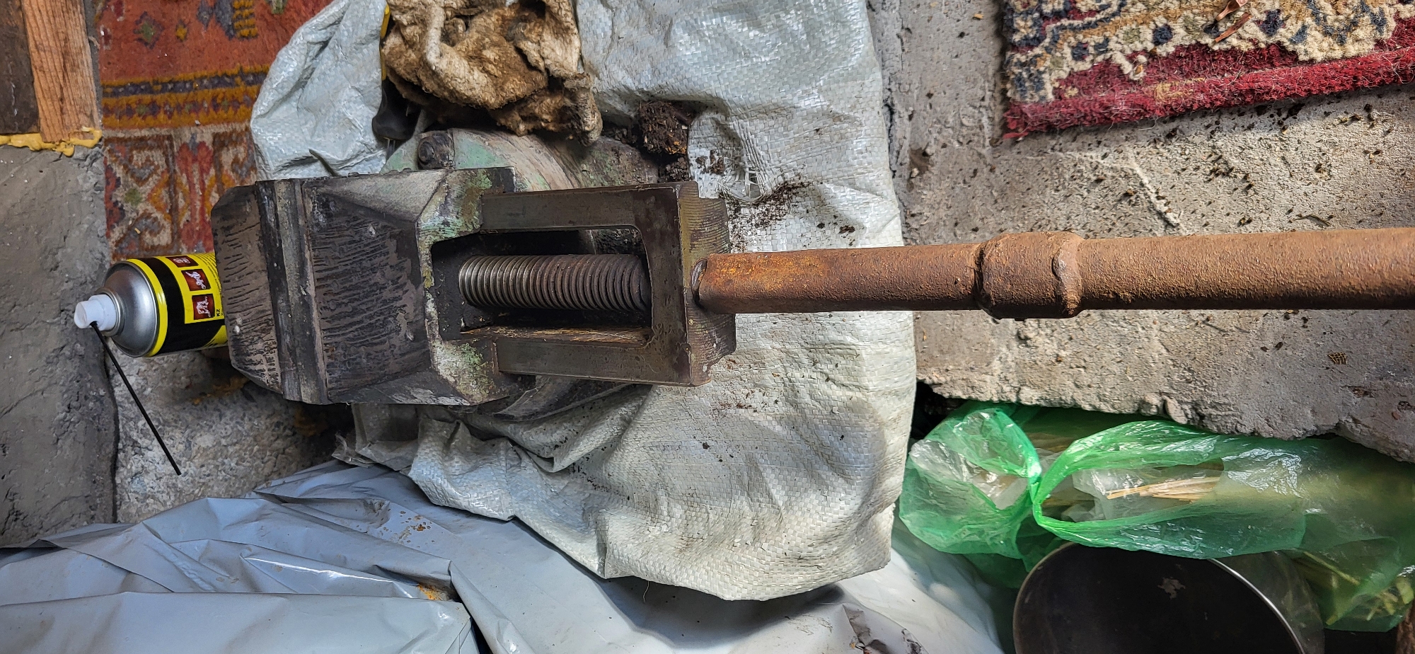 The image shows a workbench vise with a rusty metal pipe extending downwards. The vise is resting on a white woven tarp. Behind the vise, a can of spray lubricant with yellow and black labeling sits on a worn rug. To the left of the vise, another white tarp is partially visible. In the foreground, to the left and partially obscured, is a dark metal bowl and two green plastic bags containing some unidentifiable contents. The scene appears to be in a workshop or storage area, with a concrete floor and textured walls. The presence of dust and debris suggests a working environment. The location is identified as Oskemen, Kazakhstan. There are no people or interactions visible in the image.