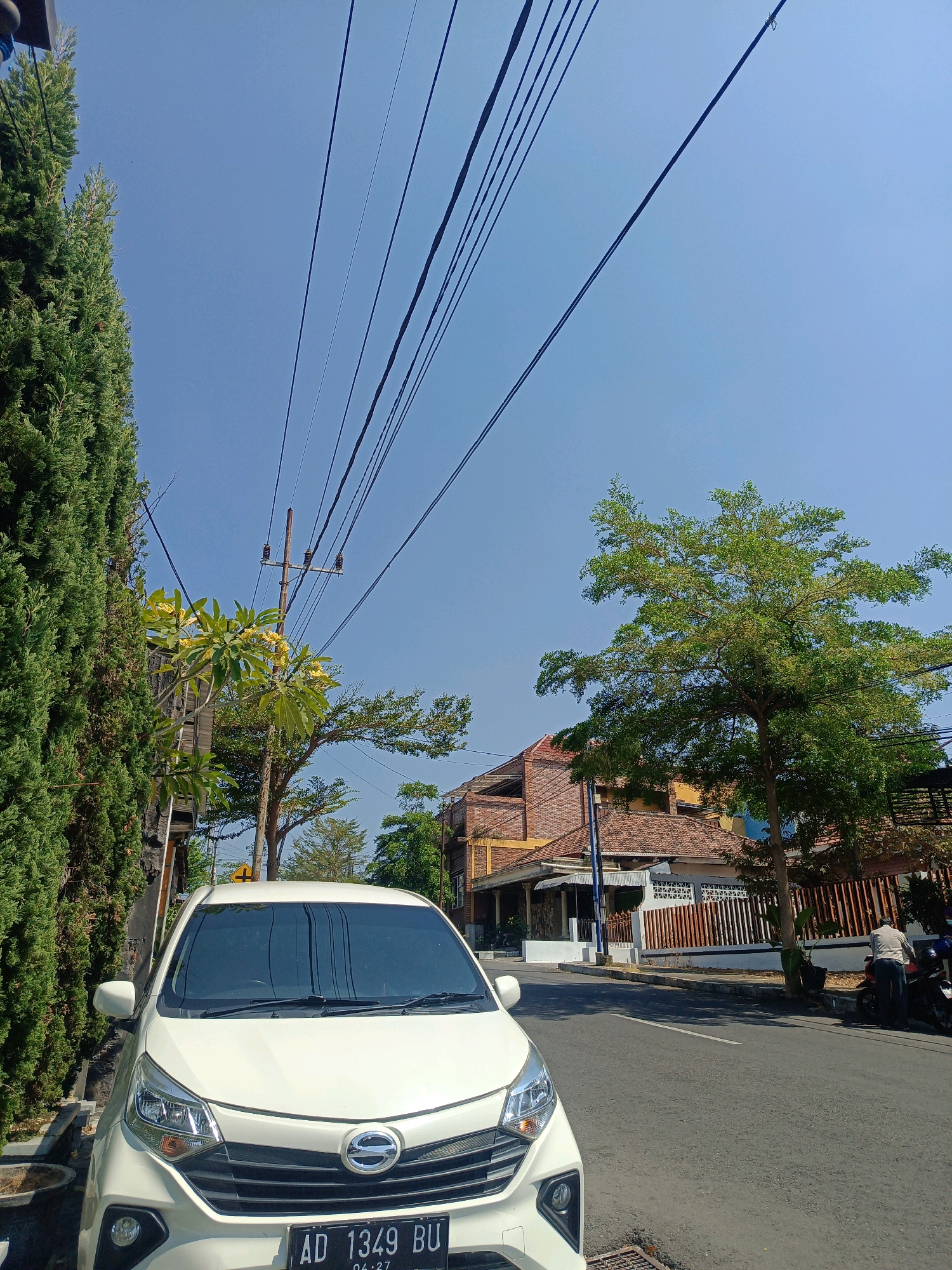 A white car is parked on the side of a road in Kepolorejo, Indonesia. The car is a compact hatchback, possibly a Daihatsu Ayla, with the license plate "AD 1349 BU". A tall, green conifer tree is to the left of the car, and further down the street, a larger, leafy tree is visible. In the background, there are houses with tiled roofs and brick walls, characteristic of Indonesian architecture. Overhead, a network of power lines stretches across the clear blue sky. A person is seen standing next to a motorcycle on the right side of the road. The overall scene depicts a typical day in a residential or semi-commercial area of an Indonesian town, with elements of both nature and urban infrastructure.