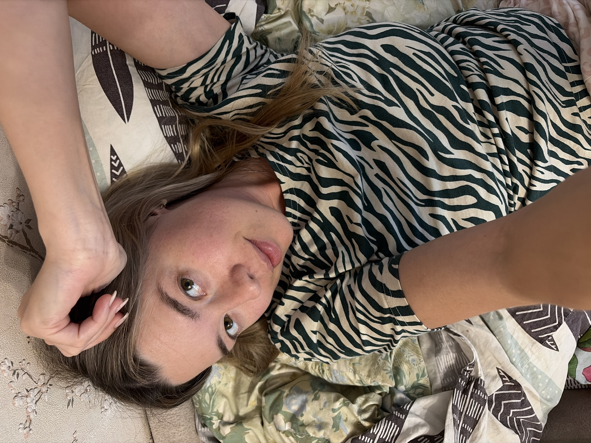 A woman lies on a bed with patterned sheets and pillowcases. She is wearing a zebra-striped top. The image appears to be taken from a close-up, slightly angled perspective. There is no information in the image indicating it was taken in Chaikovsky, Russia.