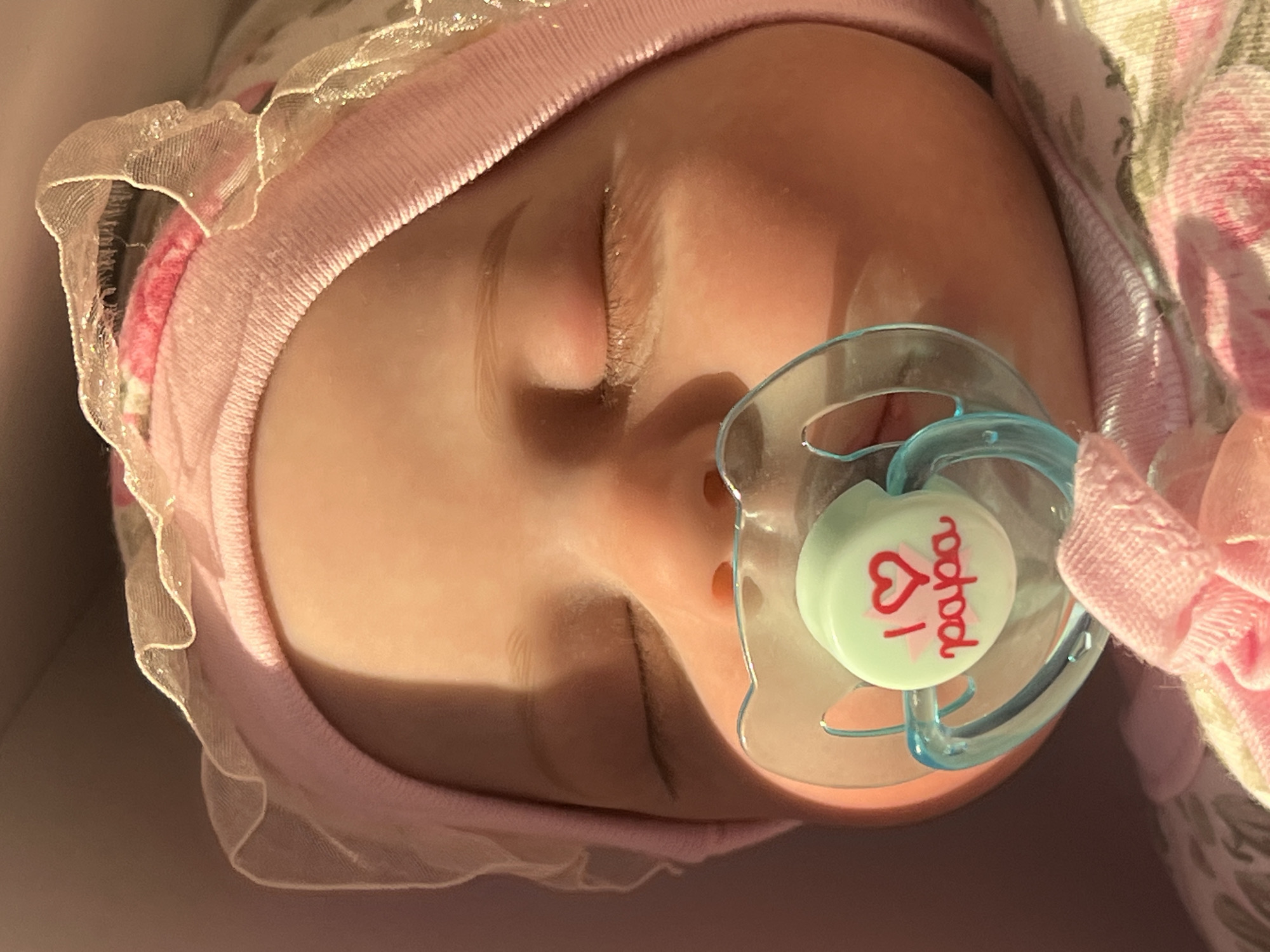 The image is a close-up, eye-level shot of a baby's face with a pacifier in its mouth. The baby is wearing a pink bonnet with lace trim. The pacifier is light green with a red heart and the word "papa" written in red. The baby's eyes are closed, and its skin is smooth and rosy. The background is a soft, out-of-focus pink and brown. There is no identifiable information in the image that could place it in Votkinsk, Russia.