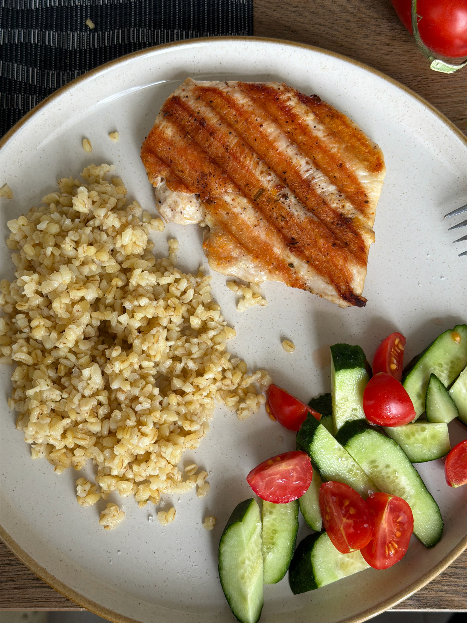 A close-up, overhead shot shows a meal on a plate. The meal consists of a grilled chicken breast with grill marks, a pile of bulgur, and a salad of sliced cucumbers and cherry tomatoes. The plate is a light beige color with a darker rim. The background is a dark patterned placemat and a wooden surface. A red tomato is partially visible in the upper right corner. There are no people or actions depicted in the image. The image was taken in the city of Khmelnytskyi, Ukraine.