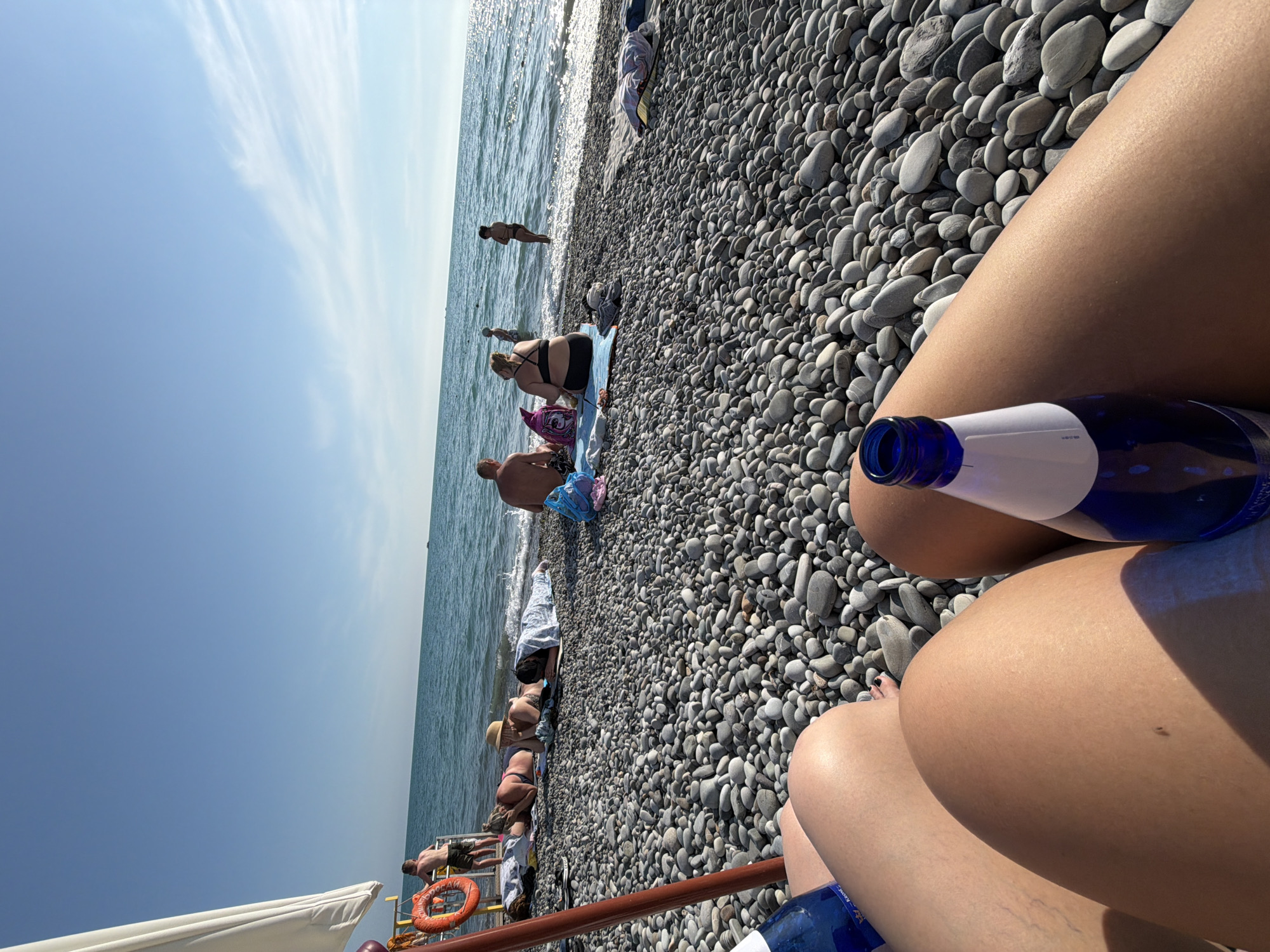 This is a first-person perspective, eye-level shot of a pebble beach in Mamedova Shchel', Russia. The foreground is dominated by the legs of two individuals, partially obscured by shadow, with two blue glass bottles positioned near their legs. The beach is covered in smooth, grey pebbles of varying sizes, extending towards the ocean. In the middle ground, several people are visible on the beach and in the shallow water. Some are lying on towels, facing away from the camera, while others are seated or standing. A life preserver ring is mounted on a pole to the left of the frame, near the water's edge. The ocean is calm with gentle waves, and the sky is a clear, bright blue with a few wispy white clouds. The sun appears to be high in the sky, casting shadows and creating a bright, summery atmosphere.