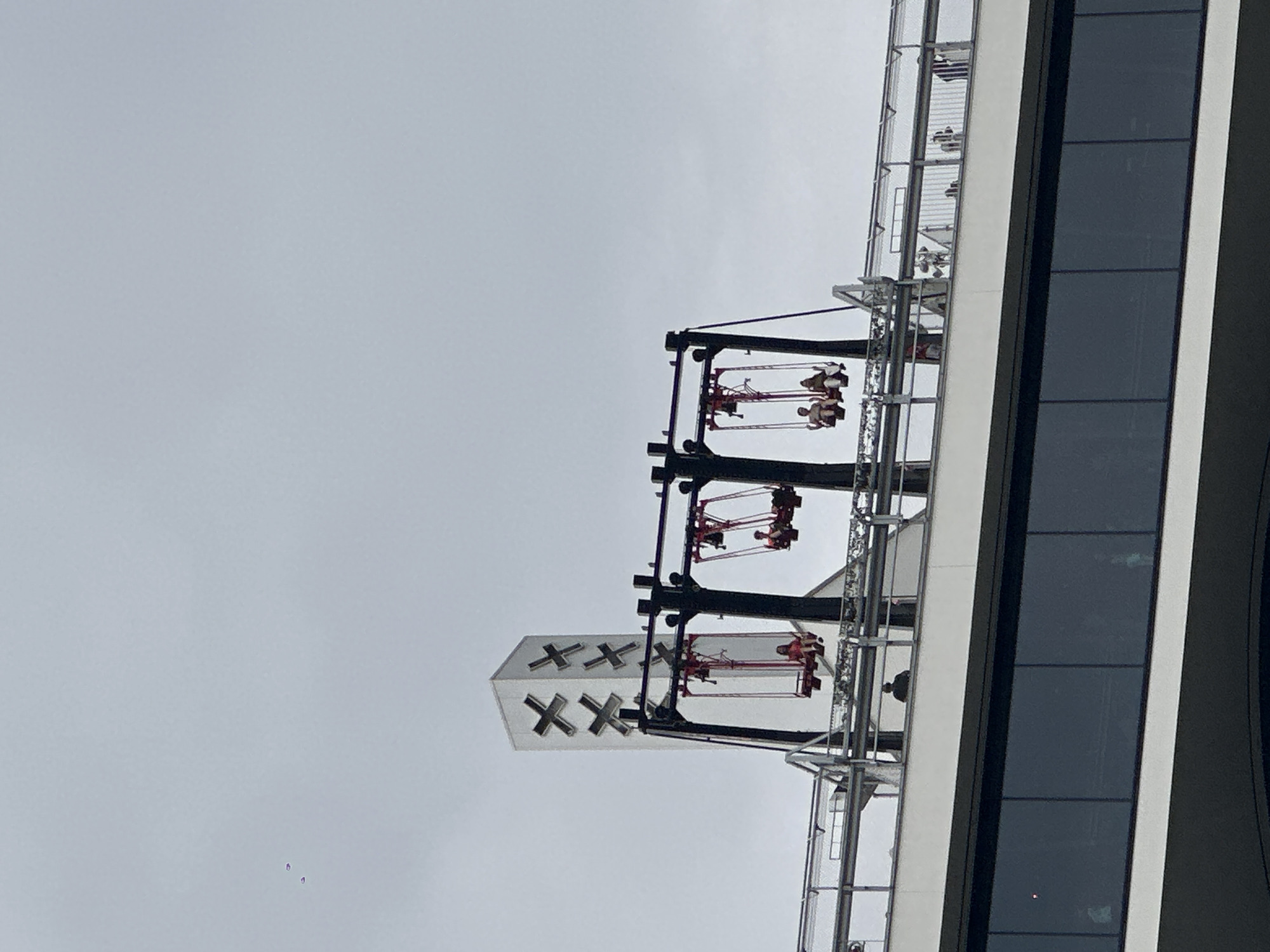 This image depicts the "Over The Edge" swing attraction at the A'DAM Lookout in Amsterdam, Netherlands. The attraction is situated atop the A'DAM Tower, a prominent skyscraper in the city. The swing is suspended from the edge of the tower, allowing riders to experience a thrilling ride with panoramic views of Amsterdam. In the foreground, a modern building with large, dark windows is visible, suggesting the perspective is from a nearby structure or the ground level. The sky is overcast, providing a diffused light for the scene. The swing itself is red and has multiple pods, each occupied by one or two individuals enjoying the ride. The iconic "XX" symbol, which is part of the A'DAM Tower's branding, is visible on the tower's structure in the background. The overall impression is an urban landscape featuring a unique tourist attraction that offers an exhilarating experience.