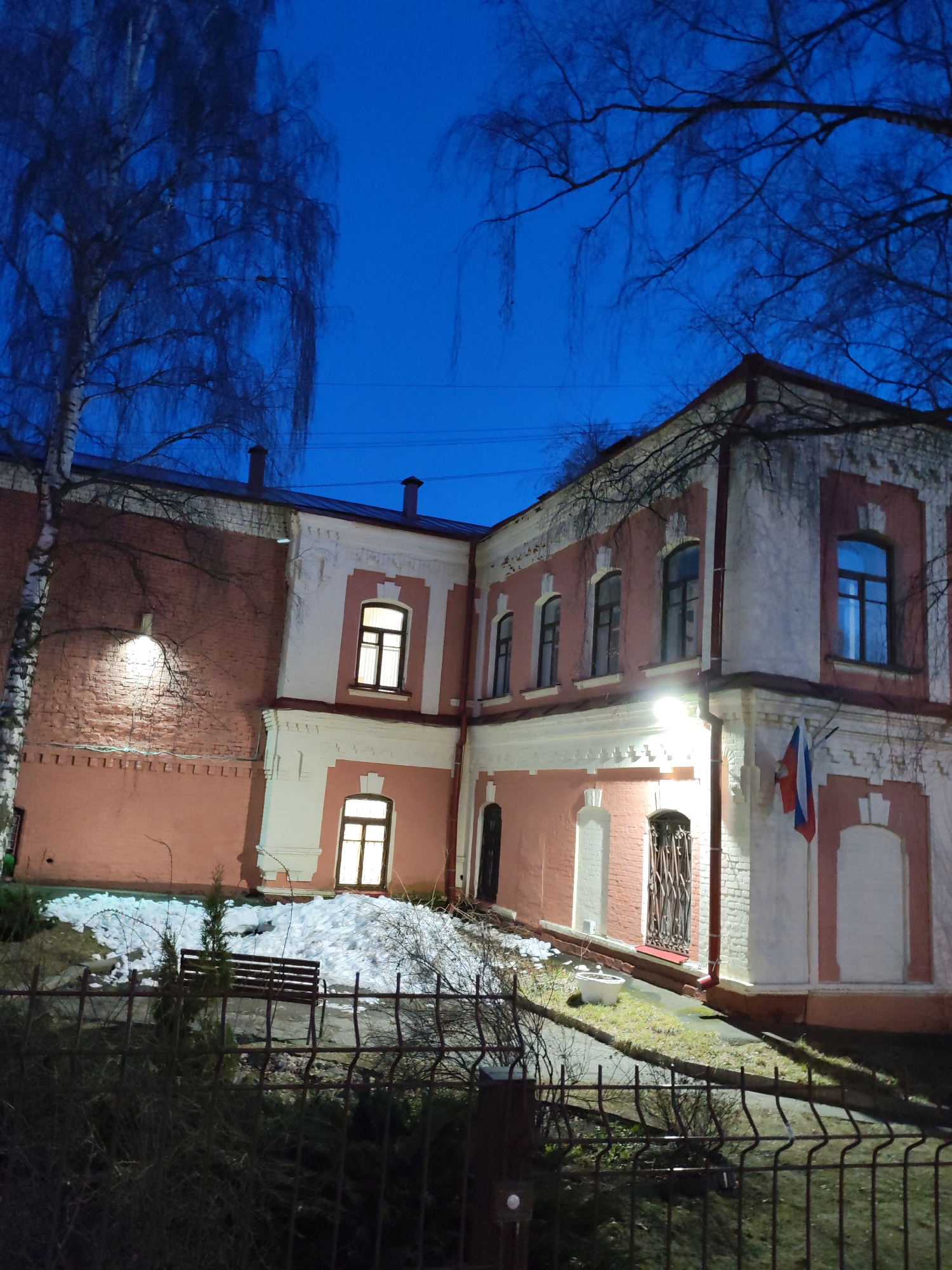 This evening photograph presents an L-shaped two-story building in Izhevsk, Russia, under a deep blue twilight sky. The architecture features reddish-pink plastered walls with striking white trim outlining windows, corners, and cornices, a style often found in older Russian buildings. Warm light emanates from several interior windows, contrasting with the cool blue of the sky and illuminating sections of the dark facade. Exterior wall-mounted lights further brighten the building.
To the left, a tall, bare birch tree stands against the sky, its delicate branches silhouetted. Other leafless tree branches are visible above and around the building. Patches of snow are scattered on the ground in the foreground, suggesting a late winter or early spring setting. A dark metal fence runs across the lower portion of the image, partially obscuring a wooden bench.
A small Russian flag, featuring horizontal stripes of red, blue, and white, is displayed vertically on the building's right-hand corner. An entrance below it is secured by a decorative metal gate. The dark roof features several chimneys silhouetted against the night sky. No people or specific activities are depicted, contributing to a quiet, serene atmosphere. No visible text is present in the image.