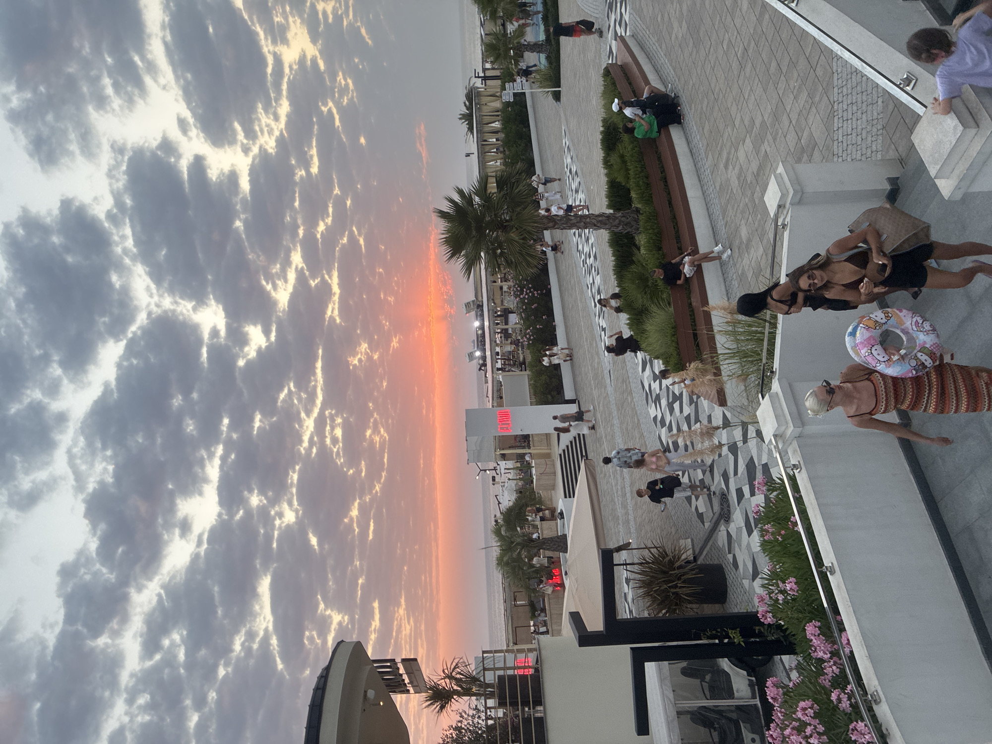 This image captures a sunset scene at a beachside promenade in Sochi, Russia. In the foreground, people are walking down a set of stairs. Two women are prominent, one wearing a patterned dress and sunglasses, carrying a bag, and the other in a bikini and shorts, also carrying a bag and a colorful inflatable ring with Hello Kitty characters. Further up the stairs, a young person in a purple shirt is visible.
The promenade features patterned paving stones, planters with pink flowers, and seating areas with people relaxing. Palm trees are scattered throughout the area, and a white structure with a red illuminated sign that reads "OLYMPIA" is visible. In the background, the sea stretches to the horizon under a dramatic sky filled with textured clouds tinged with the orange and pink hues of sunset. Beach umbrellas and structures are set up along the coastline, indicating a popular resort area. The overall atmosphere appears to be one of leisure and evening relaxation.