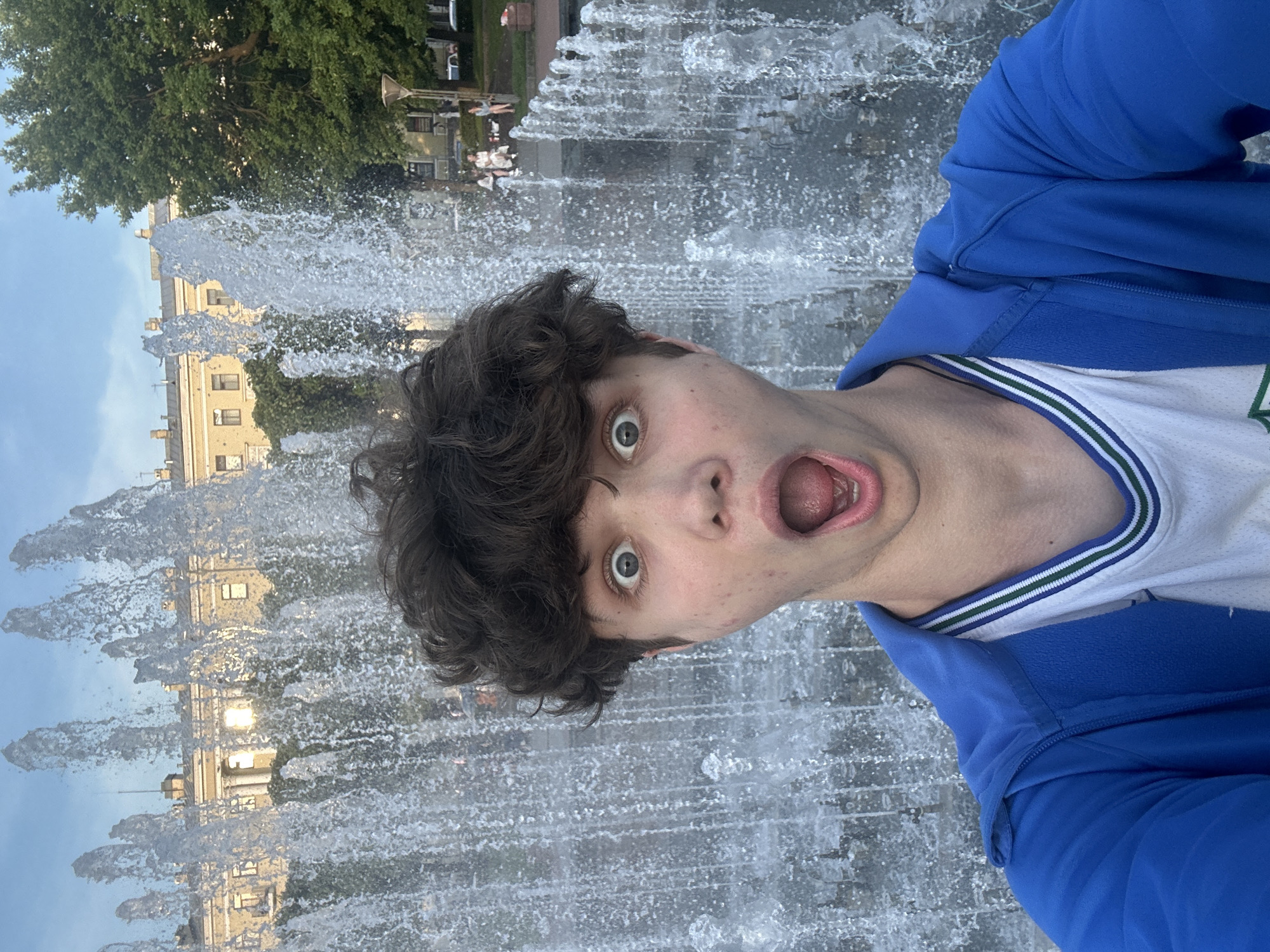 A young male with messy brown hair and wide, surprised eyes takes a selfie in front of a tall water fountain in what appears to be a city park. He is wearing a bright blue zipped jacket over a white t-shirt with green and white trim around the collar. His mouth is open in an expression of shock or excitement. Behind him, the water from the fountain creates a curtain of droplets. In the background, a classical-style building with a light-colored facade and multiple windows is visible, as well as lush green trees and other park-goers. The sky above is partly cloudy and appears to be late afternoon or early evening.