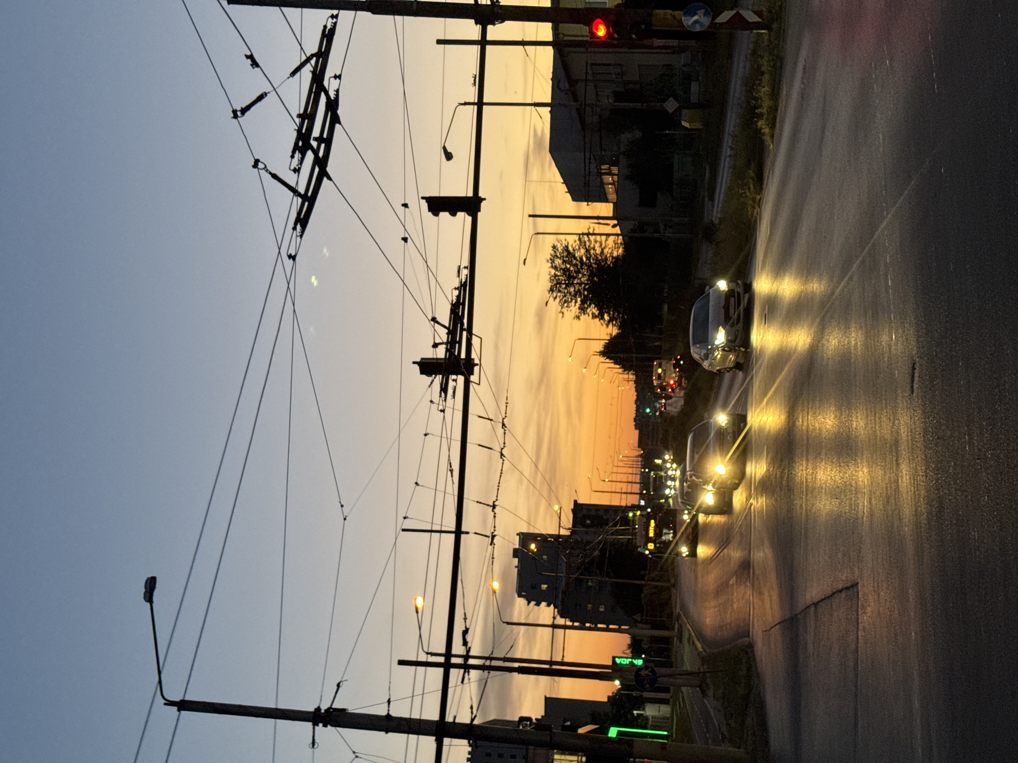 The image captures a street scene in Varna, Bulgaria, at dusk. The sky displays a gradient of colors, transitioning from a pale blue at the top to warm orange and yellow hues near the horizon, indicating the setting sun. Overhead, a complex network of power lines and trolleybus wires crisscrosses the frame, supported by utility poles. Streetlights are illuminated, casting a yellow glow on the wet asphalt road. Several cars are visible, their headlights on, reflecting on the road surface. One car, a white sedan, is in the foreground, moving towards the right. In the middle ground, a bus is seen, and other vehicles are in motion further down the road. Buildings line the street, some of which are silhouetted against the sky. A traffic light shows a red signal on the right side of the image. The overall impression is of an urban environment at the end of the day, with traffic and infrastructure elements dominating the scene.