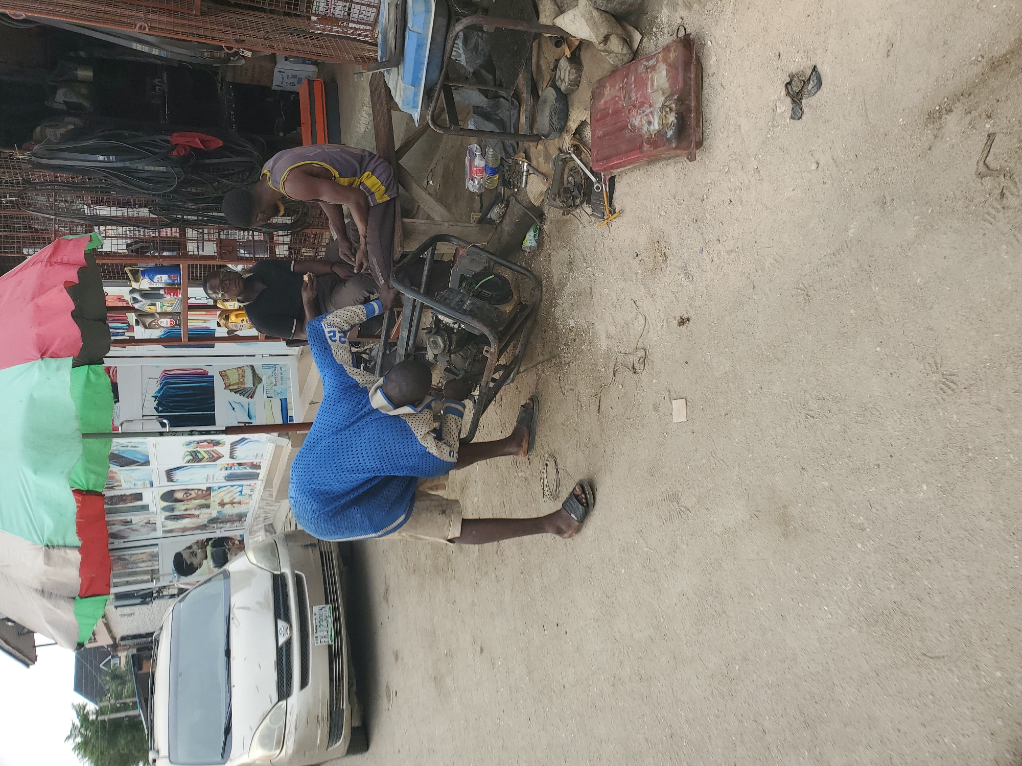 This image captures an outdoor scene in Lagos, Nigeria, depicting a generator repair operation during daylight hours.
In the foreground, a sandy-colored ground is scattered with various tools, including wrenches and a hammer, along with generator components like a red fuel tank and plastic bottles, some containing "PURE WATER".
Three men are actively involved in or observing the repair work. One man, wearing a blue and white patterned long-sleeved top with the number "22" on it and light-colored shorts, is squatting and working on a generator frame. He is wearing black flip-flops. Another man, dressed in a dark grey sleeveless top and dark trousers, is bent over an adjacent generator, also engaged in repairs. A third man, wearing a black shirt and glasses, is seated nearby, looking towards the activity.
Multiple generators and their disassembled parts are central to the scene. To the left, a large, multicolored umbrella (red, green, and light blue sections) provides shade. Partially visible behind the umbrella is a light-colored van, likely a Toyota Hiace, with a Nigerian license plate clearly showing "LAGOS KJJ 238", confirming the location.
The background reveals a roadside shop or stall. Shelves display various items, including multiple bottles of engine oil, some labeled as such. Further back, there are stacks of belts or tires, and a display case with what appears to be textiles or household goods. A metal security grille is also visible, common in commercial settings. The overall impression is a bustling, practical work environment.