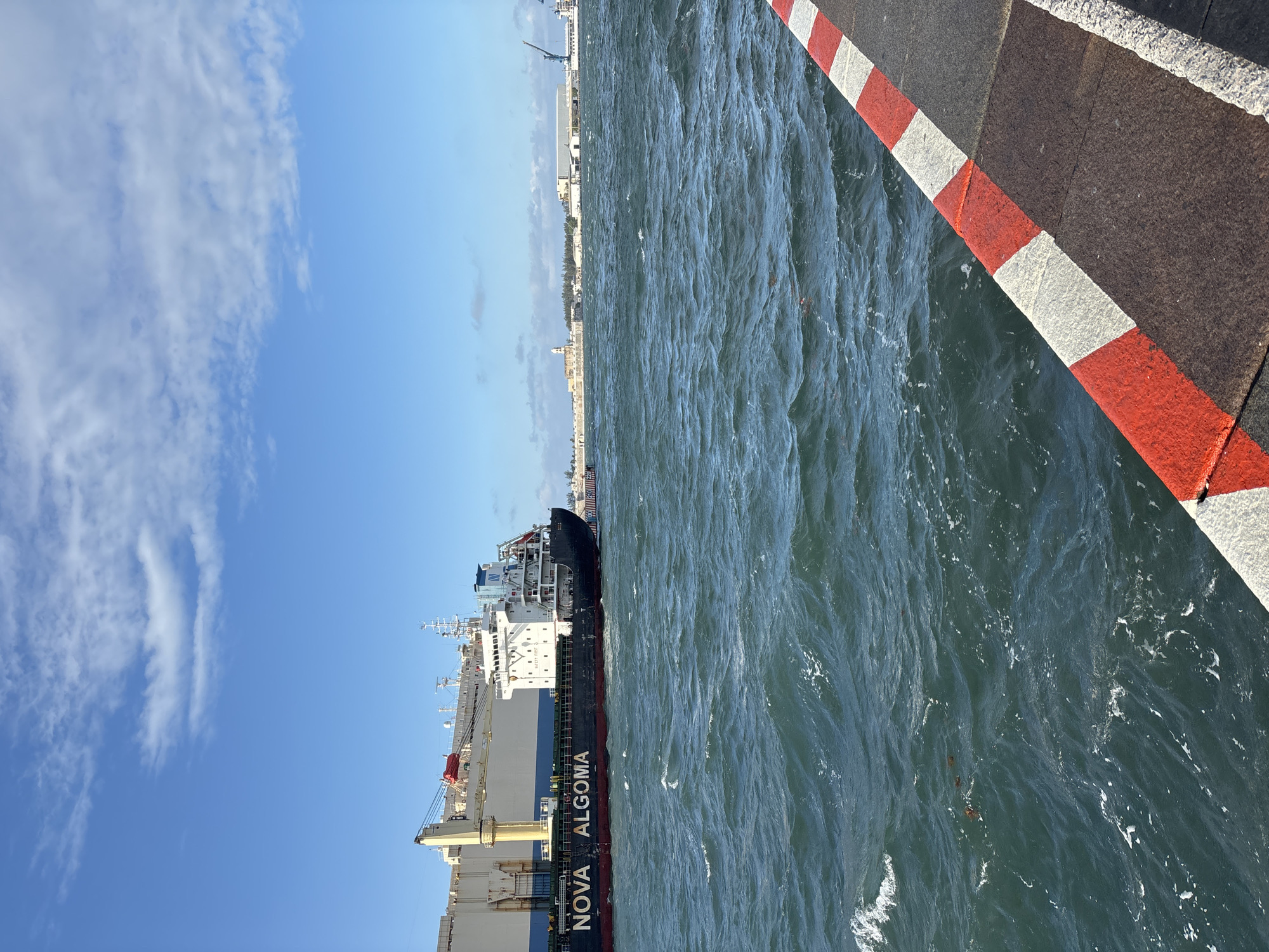 The image captures a large cargo ship, "NOVA ALGOMA", docked along a pier in an urban setting. The pier's edge is marked with red and white stripes. To the right, the scene consists of the water of the port. The background shows a cityscape under a clear blue sky with some clouds.