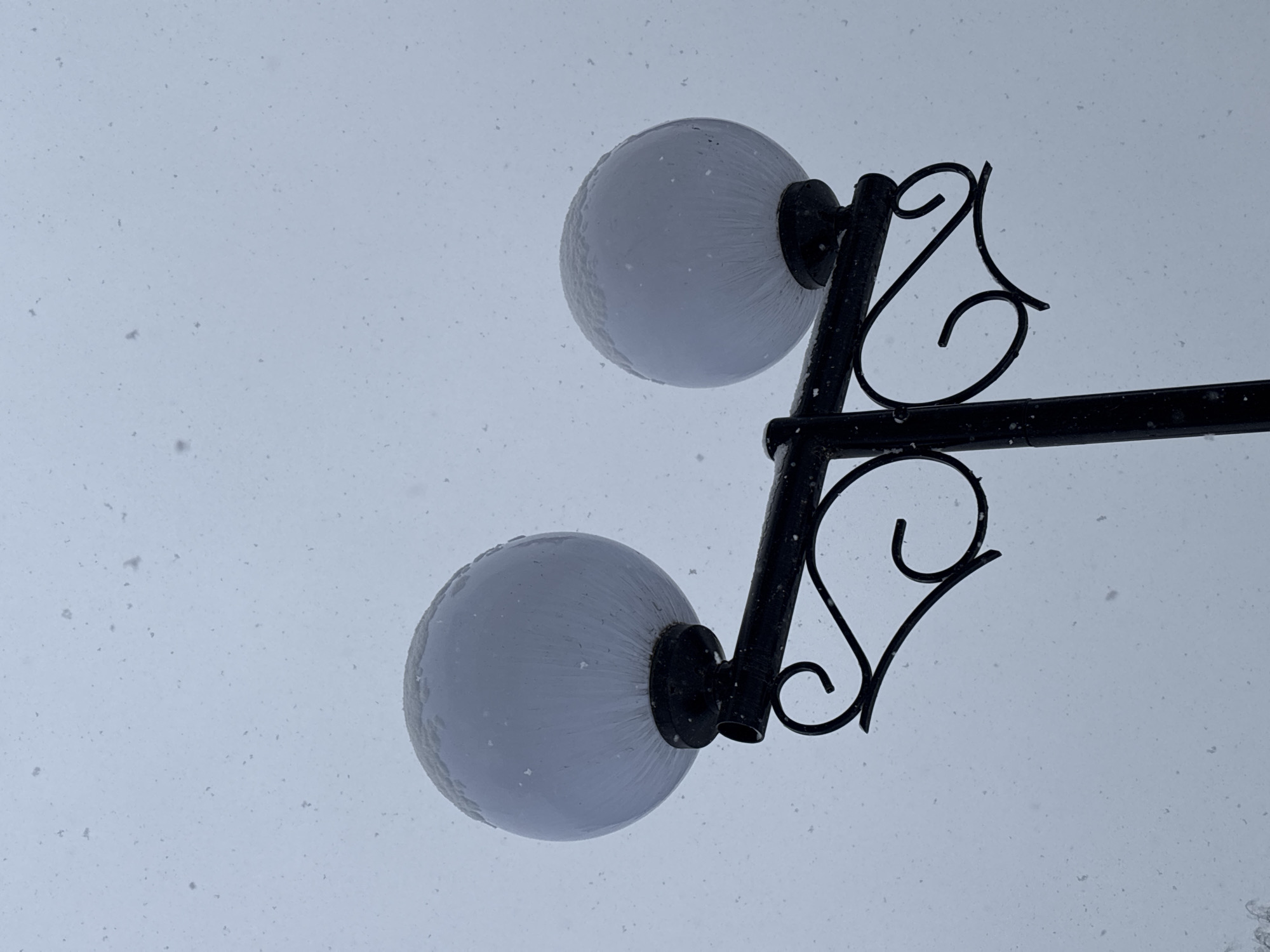 A low-angle shot depicts a black metal lamp post with two frosted spherical globes, presumably in Chaikovsky, Russia. The sky is a pale gray, and snow or light debris is visible, possibly falling. The lamp post features decorative wrought iron elements. The globes appear to be illuminated, although dimly, and have a slight buildup of snow or frost on their surfaces.