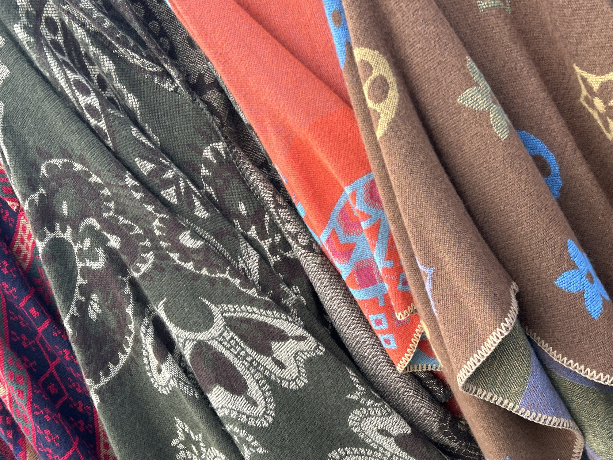 The image shows a close-up, top-down view of several folded textiles, appearing to be scarves or blankets, layered on top of each other. The topmost textile has a dark blue and red intricate geometric pattern. Beneath it is a dark green textile with a repeating paisley-like pattern in a lighter shade of green and white. The next visible textile is a vibrant orange with a geometric pattern featuring pink and blue squares and diamond shapes. The bottommost textile is a brown fabric with repeating light blue and yellow floral or abstract motifs, and a visible stitched hemline. The lighting is diffused, and the textures of the fabrics are clearly visible, indicating a soft, possibly wool or acrylic material. The image does not contain any information or elements that would allow for the determination of its location in Mtskheta, Georgia.