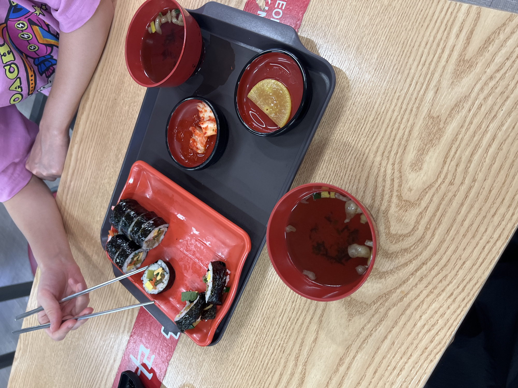 This media file captures a top-down view of a meal on a light-colored wooden table. The scene includes a black tray holding several red bowls and a red rectangular plate. On the tray, there are two small red bowls, one containing kimchi and the other a slice of pickled radish. A larger red bowl with broth and some garnishes sits at the top left of the tray.
On the red rectangular plate, there are several pieces of kimbap, a Korean dish similar to sushi rolls. A person's arm and hand, wearing a pink t-shirt with a graphic design of a rocket and planets, are visible on the left side of the image, holding a pair of silver chopsticks. The chopsticks are positioned above the kimbap, suggesting the person is about to eat. Another red bowl filled with broth and garnishes is placed to the right of the tray.
A red and white striped tape with Korean text is visible on the tray and extending onto the table. The visible text includes "EOI". The overall impression is of a casual meal, possibly at a restaurant in Seoul, South Korea, given the context. The lighting suggests it is daytime. The focus is on the food and the act of preparing to eat.