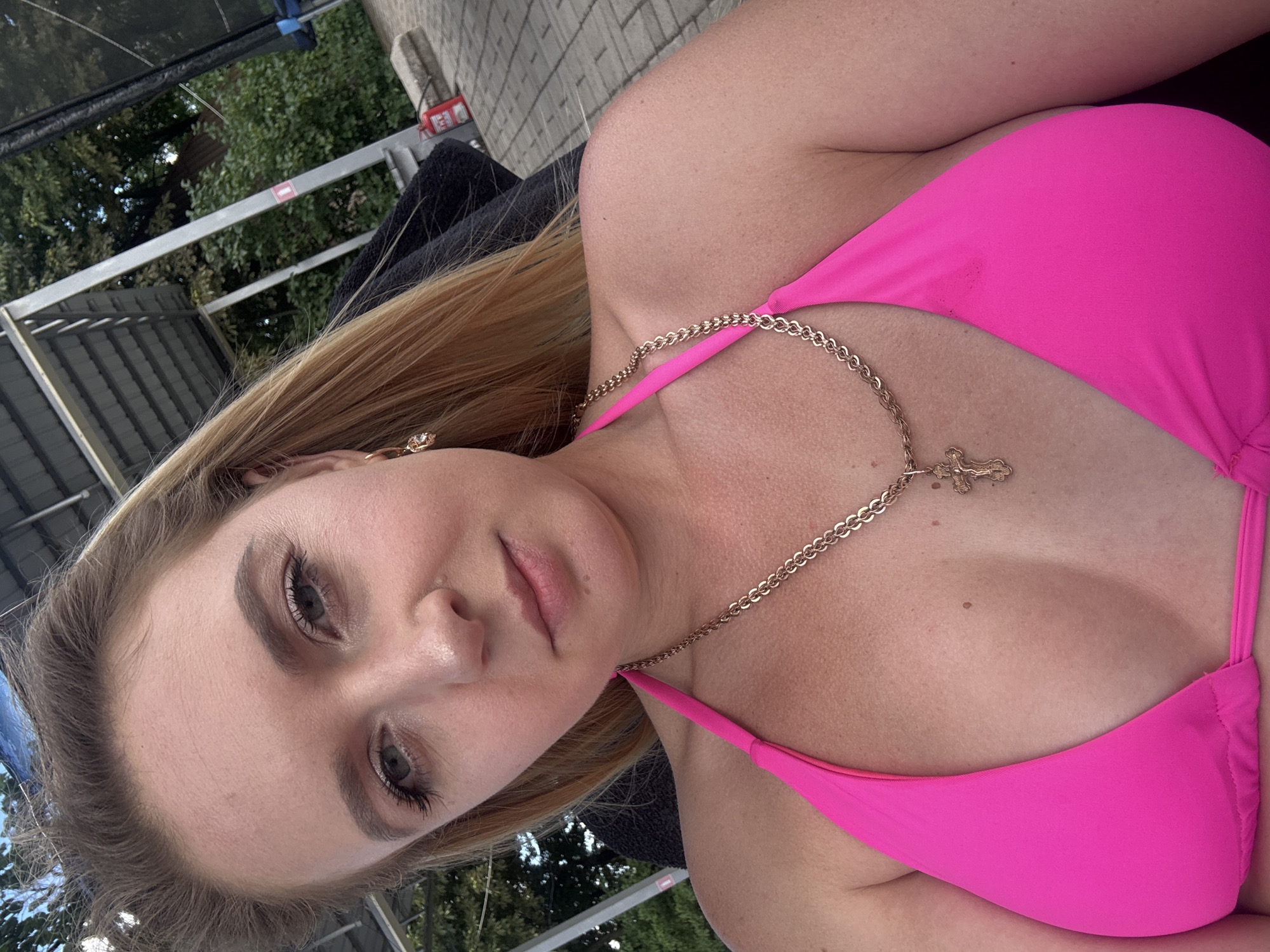 This is a close-up, vertically oriented selfie of a young woman lying down outdoors. She is wearing a bright pink bikini top with a gold chain necklace featuring a cross pendant. Her blonde hair is visible, and she has on makeup. The background shows trees and a tiled surface, suggesting an outdoor leisure setting. The lighting indicates it is daytime.
The image appears to have been taken in Makiivka, Ukraine.