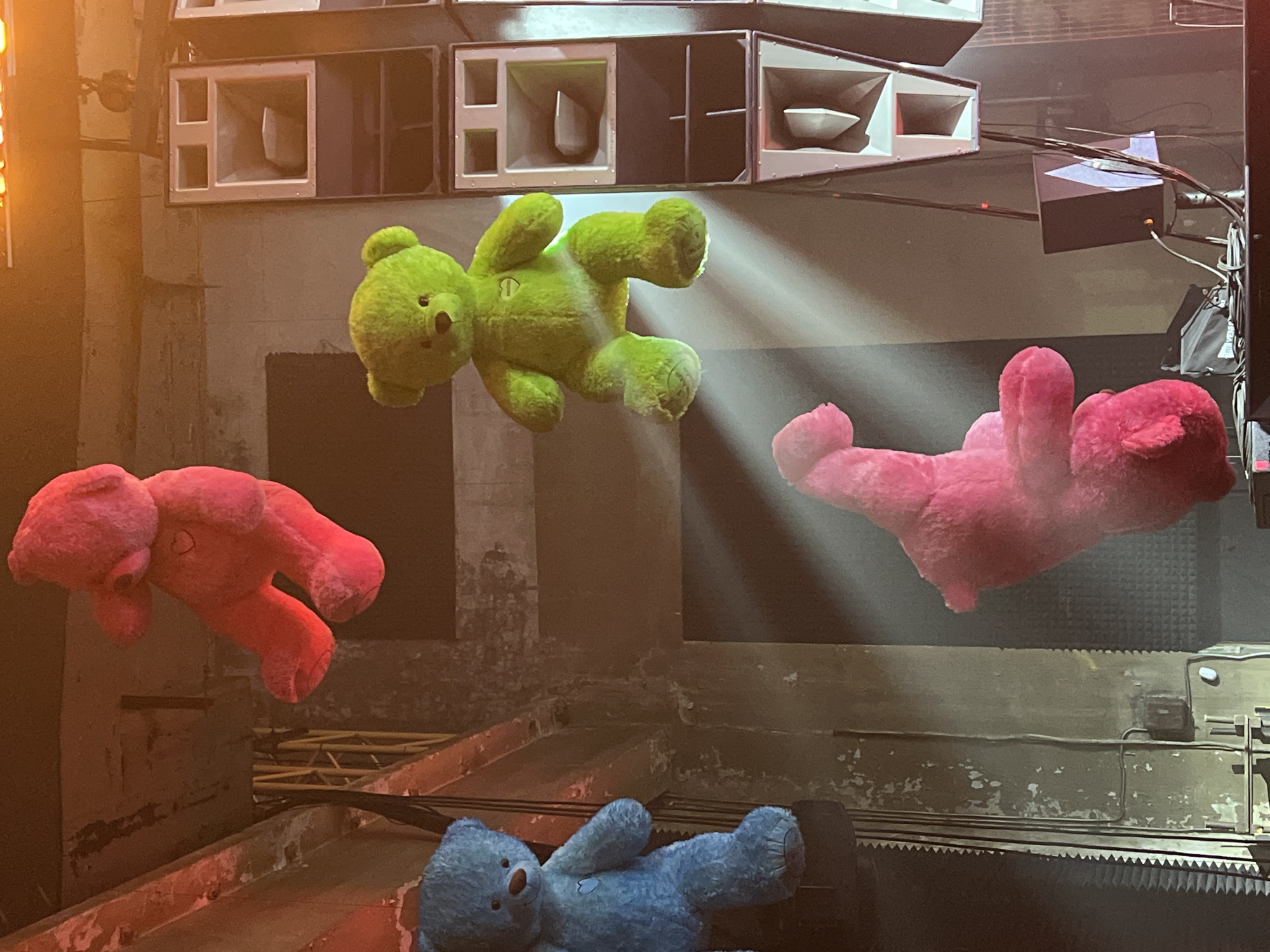 The image depicts a surreal indoor scene featuring several plush teddy bears set against an industrial backdrop, likely in Moscow, Russia.
**Subjects:** The primary subjects are four teddy bears: a large, vibrant green teddy bear suspended horizontally in the upper center, two pink teddy bears floating on either side of the green one (one on the left facing right, one on the right facing left), and a blue teddy bear partially visible lying on a dark surface in the lower foreground. The green teddy bear is illuminated by a strong diagonal spotlight.
**Setting and Objects:** Above the bears, a stack of large, horn-loaded audio speakers, typical of club or concert sound systems, dominates the background. These speakers have dark frames and some feature white or grey interiors. The walls and structures behind the bears and speakers appear to be made of concrete or an industrial material, showing signs of wear, grime, and possibly peeling paint. Cables and wires are visibly running across the walls and connecting to the speakers. A dark, rectangular object with purple tape is suspended near the upper right. The surface in the foreground below the bears has a dark, corrugated, or jagged edge, suggesting an industrial platform or stage.
**Activity/Event:** The arrangement of the suspended teddy bears within this industrial, speaker-laden environment suggests an art installation, a unique decorative element for a club, party, or event. The dramatic lighting on the green bear enhances this impression.
**Notable Details:** The scene is dimly lit, with a powerful spotlight creating distinct light rays that highlight the green bear, implying a nighttime or dark indoor setting. The juxtaposition of the soft, playful teddy bears with the harsh, utilitarian industrial setting and massive sound system creates a striking and somewhat whimsical atmosphere. No discernible text is visible in the image.