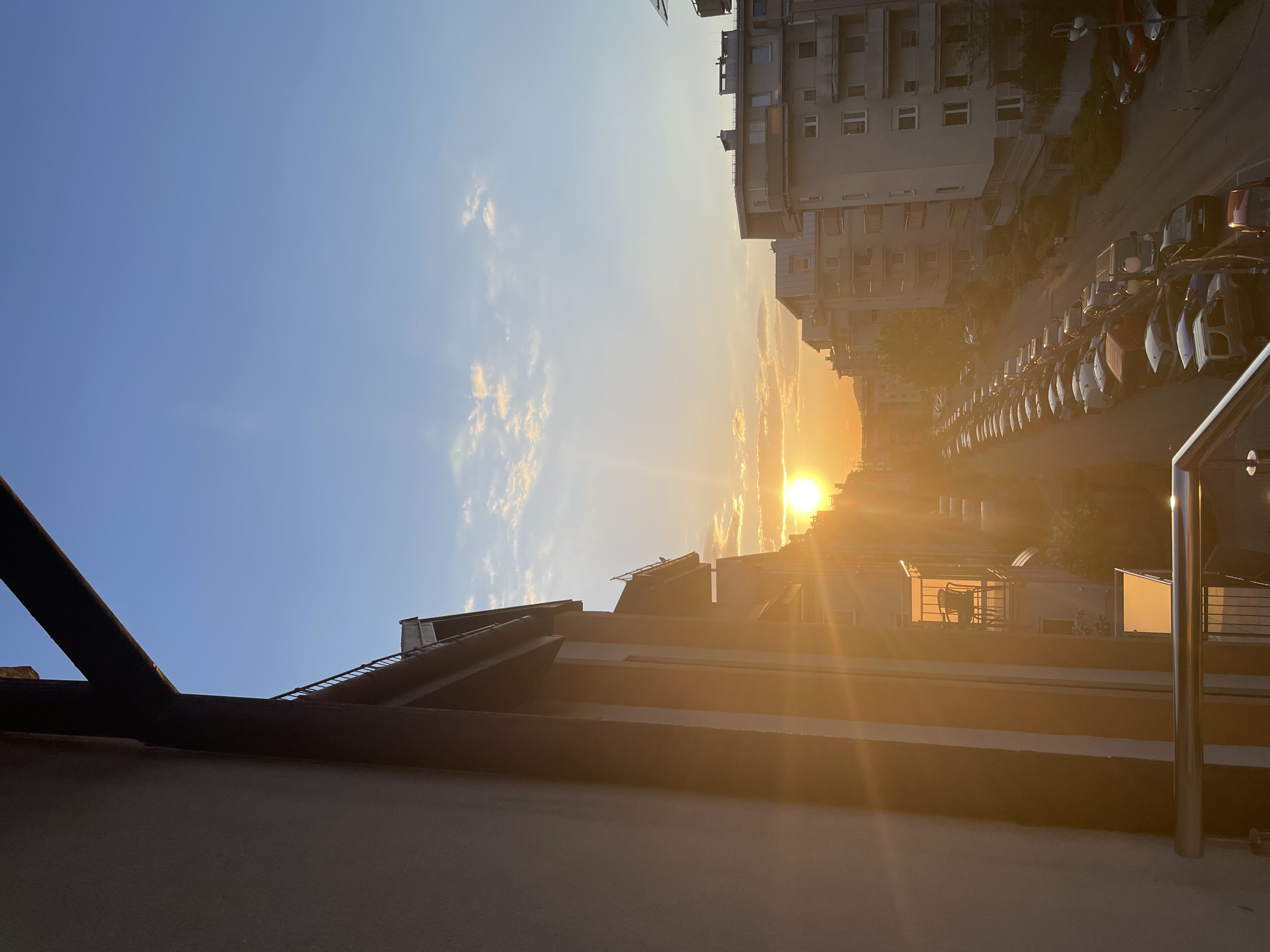 The image shows an urban cityscape at sunset. The view is from a high vantage point, possibly a balcony or rooftop, overlooking buildings and a street. The sun is setting on the horizon, casting a golden light across the scene. A row of parked cars lines the street next to a tall building. The sky is blue with some clouds near the horizon. There are no people visible in the image. The location is Unknown, Unknown.