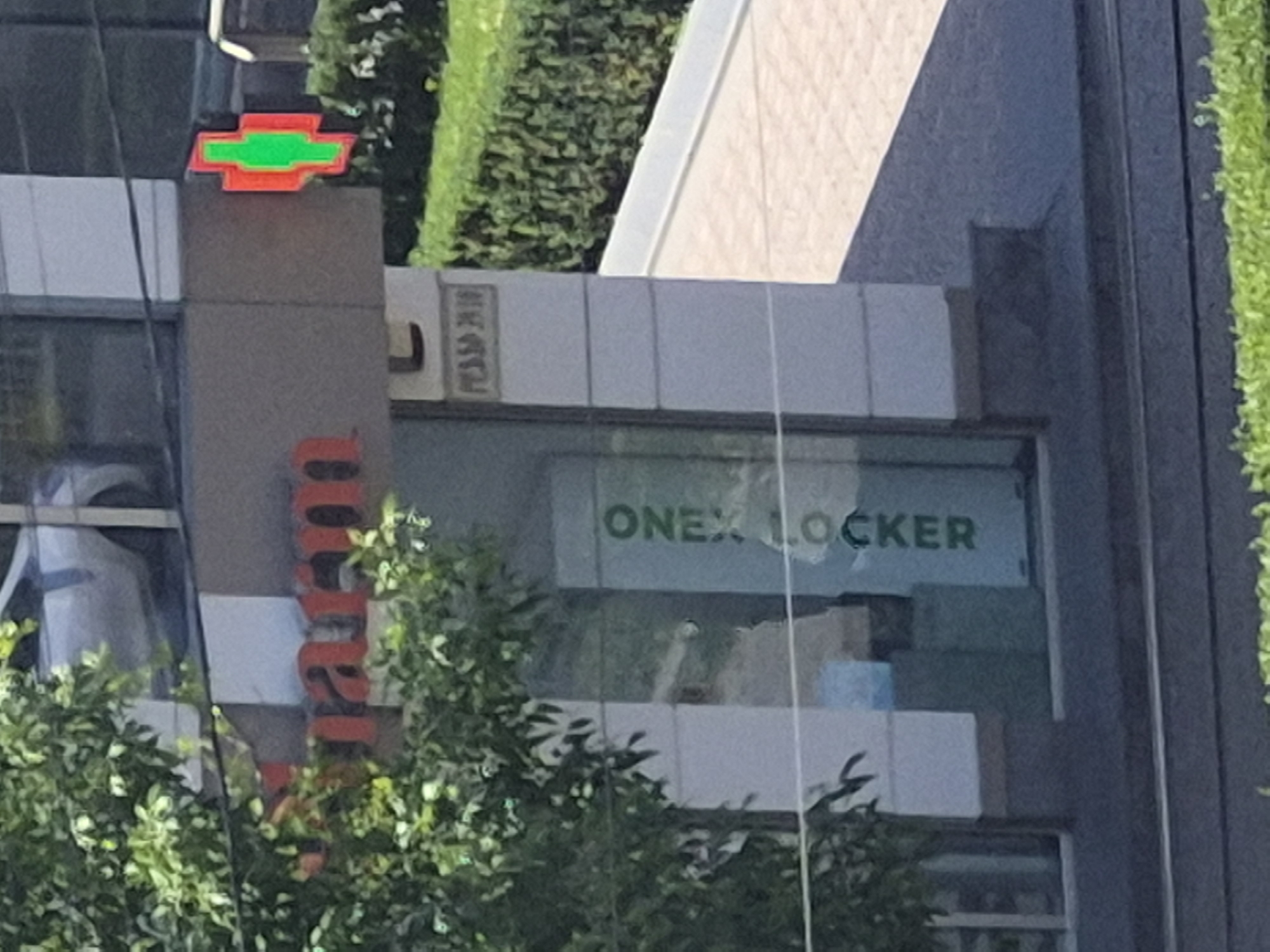 An eye-level shot captures a section of a modern building in Yerevan, Armenia, bathed in bright daylight. The building features large glass windows and grey paneling, with lush green foliage from trees or hedges framing parts of the structure.
A prominent red and green Chevrolet logo is affixed to the upper part of a grey pillar on the building's exterior. Below and to the left of the Chevrolet logo, a vertical red sign displays the letters "amaran," indicating a business name or brand. To the right of the Chevrolet logo, on a white panel, faint Cyrillic text appears to read "РЕД 30" (RED 30).
Through a large window, the words "ONEX LOCKER" are clearly visible in green lettering, indicating a business or service located inside. Various boxes and items are faintly discernible behind the "ONEX LOCKER" sign, suggesting storage or package handling activities. The scene is bright, implying a clear, sunny day, with reflections visible on the glass surfaces of the building. No people are visible, and the scene primarily focuses on the building's exterior details and signage.