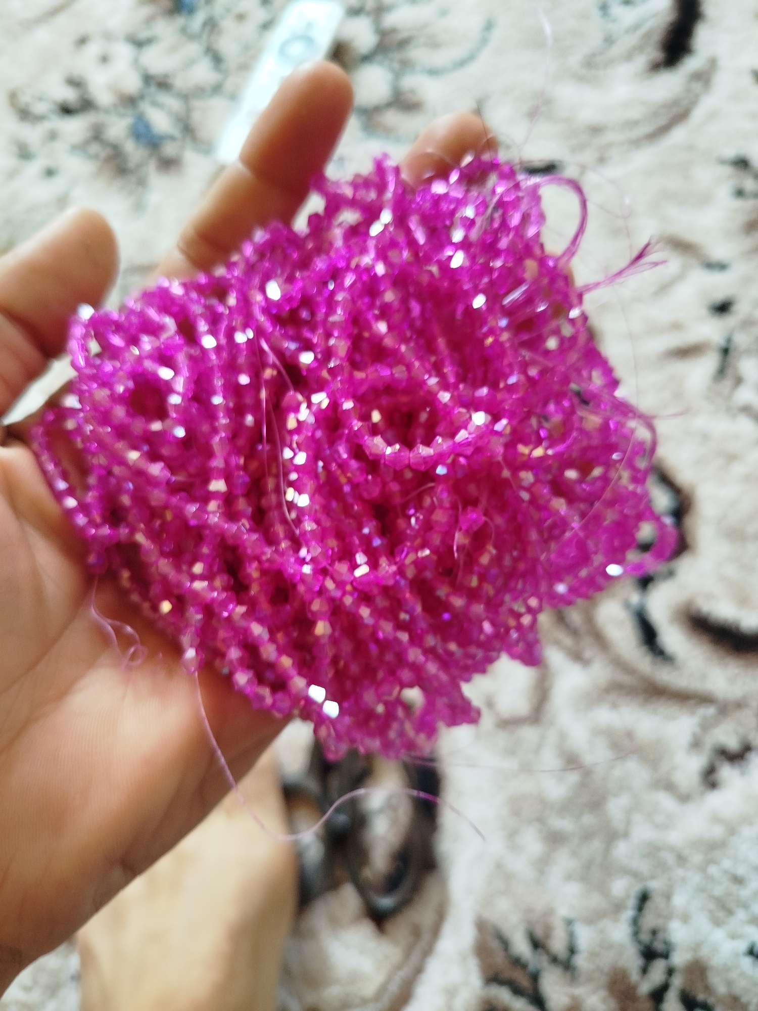 A hand holds a cluster of small, bright pink, faceted beads. The beads are strung together on thin, translucent threads, creating a textured, somewhat amorphous shape. The hand is open, with fingers slightly curled, supporting the beaded mass. The background is a soft-focus carpet with a muted pattern of beige, white, and dark lines. A small portion of another hand or foot is visible at the bottom of the frame. The lighting is bright, causing highlights to gleam on the beads. The overall impression is a close-up, intimate shot of a craft material. The location is not discernible from the image itself, and therefore cannot be placed in Kokand City, Uzbekistan.