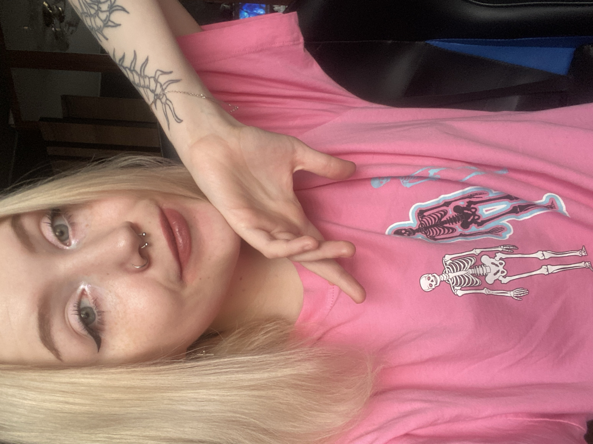 A close-up, chest-up portrait shows a young blonde female in an indoor setting in Izhevsk, Russia. The subject has fair skin with visible freckles, light-colored eyes with winged eyeliner and light eyeshadow, and lips with a brownish-pink gloss. She wears a septum piercing, a horseshoe-shaped ring, and small stud earrings. Her long, straight blonde hair frames her face. She is wearing a pink t-shirt featuring screen-printed graphics of human skeletons; one skeleton is predominantly white with red eyes, and another is darker with a light blue aura. The subject's left hand is positioned with fingers under her chin or jawline. Her right forearm, partially visible, displays a tattoo resembling an arthropod or centipede and a delicate chain bracelet. The background is dark and indistinct, with a black object featuring a blue accent, consistent with a gaming chair, visible on the right side. The subject maintains a direct gaze towards the camera with a slight smile.