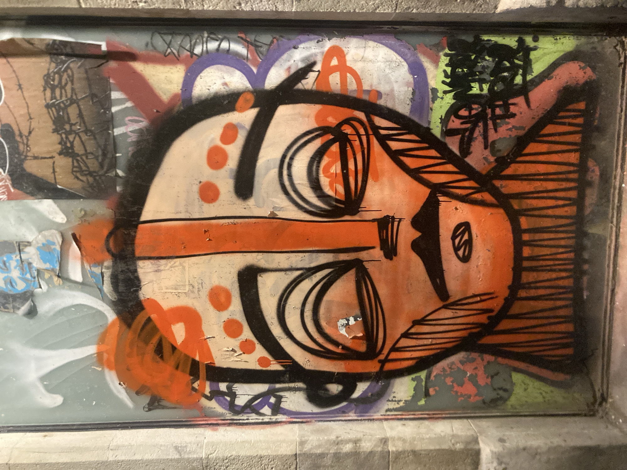 The image shows a graffiti artwork on a glass surface, likely a window or a storefront, in Barcelona, Spain. The artwork features a stylized, orange face with black outlines. The face has large, almond-shaped eyes with black outlines, a small black mustache, and a wide mouth indicated by vertical black lines. There are orange circular markings on the forehead and cheeks. The background is a collage of other graffiti tags and images, with colors including purple, green, red, and white. The surface appears weathered, with peeling paint and layers of different graffiti. The surrounding environment is not clearly visible, but the edge of a stone pillar is present on the left side of the image. The overall impression is of urban street art on a public surface.