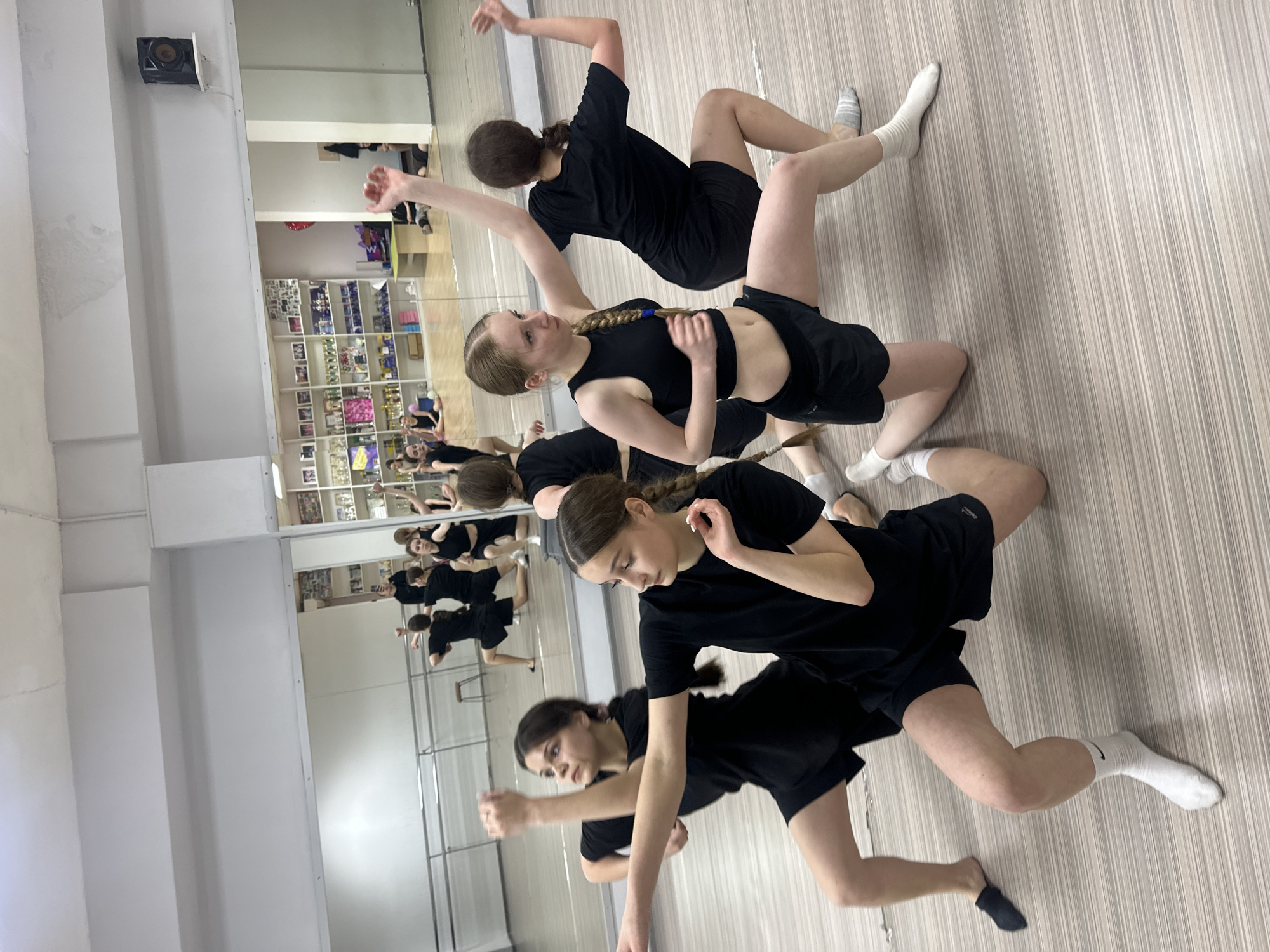 The image depicts a group of young female dancers practicing in a brightly lit dance studio, likely located in Votkinsk, Russia. The studio features light-colored wooden or laminate flooring and white walls, with a significant portion of one wall covered by large mirrors.
Five dancers are prominently visible in the foreground and midground, with their reflections, along with those of other dancers, appearing in the mirrors. All are dressed in black activewear, including t-shirts, crop tops, and shorts, and wear various white, grey, and black socks. Several dancers have braided hair. They are performing contemporary or modern dance movements, focusing on floor work and dynamic, angular poses. Their expressions convey concentration and dedication to their practice.
Reflected in the mirrors are a ballet barre running along the wall, and a shelving unit laden with trophies, framed pictures, and other items, suggesting a history of dance achievements. A speaker is mounted high on a wall near the ceiling. A white Nike swoosh logo is clearly visible on one of the white socks worn by a dancer. Other texts and a "W" logo are faintly discernible on the reflected shelves but are illegible. The overall scene indicates a focused training session within a dedicated dance environment.