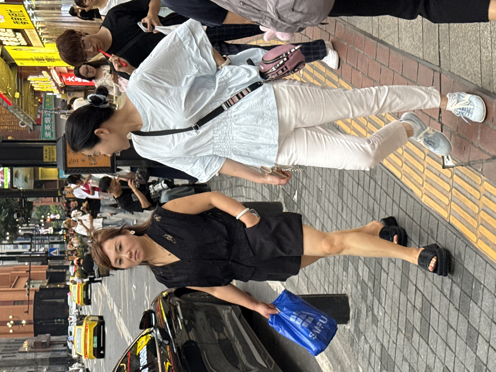 Two women are walking on a city sidewalk in Yuzhong District, China. The woman on the left is wearing a black lace sleeveless top and black shorts. She is carrying a blue plastic shopping bag in her left hand and wearing black platform sandals. She has a white bracelet on her left wrist. The woman on the right is wearing a light blue ruffled short-sleeved top and white pants. She has a dark crossbody bag with a pink pouch attached. She is wearing light blue sneakers. The sidewalk is paved with gray tiles and has a yellow tactile paving strip along the edge. In the background, there are shops with bright signs, other pedestrians, and yellow taxis on the street. A black car is parked on the left side of the image. The scene appears to be during the daytime.