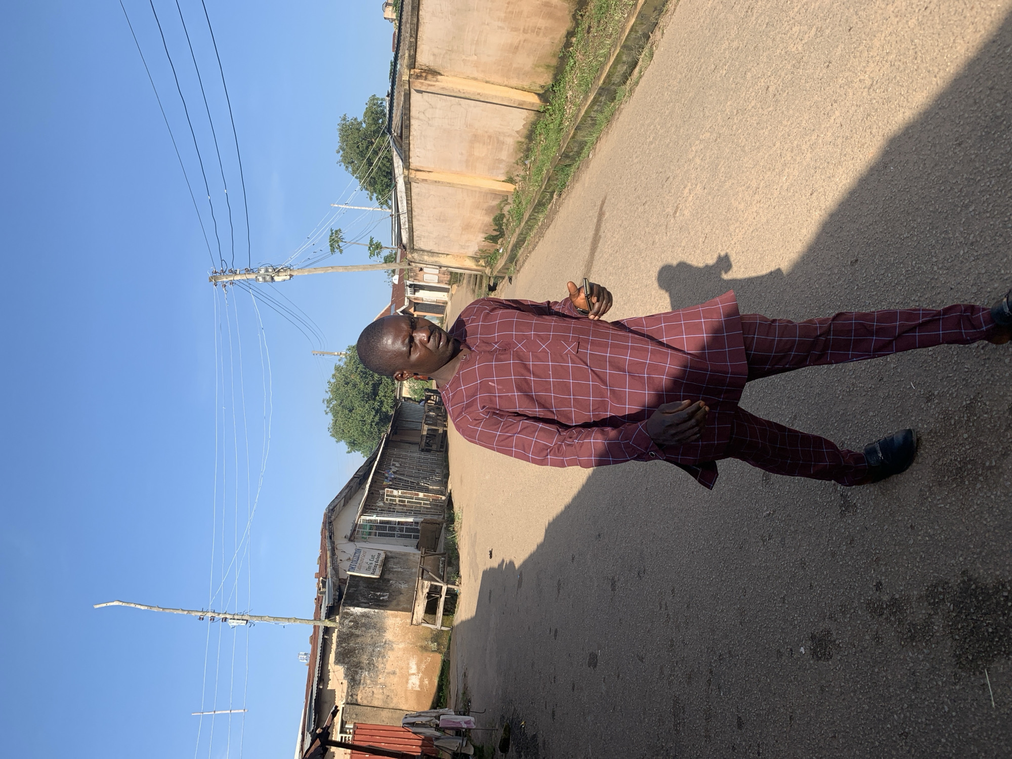 The image shows a man standing on an asphalt road in Jos, Nigeria. He is wearing a maroon and blue checkered traditional African outfit. The man is holding a cell phone in his right hand. In the background, there are buildings with corrugated metal roofs and painted walls, as well as utility poles with power lines. The sky is clear and blue. The overall impression is that of a sunny day in an urban or semi-urban environment.