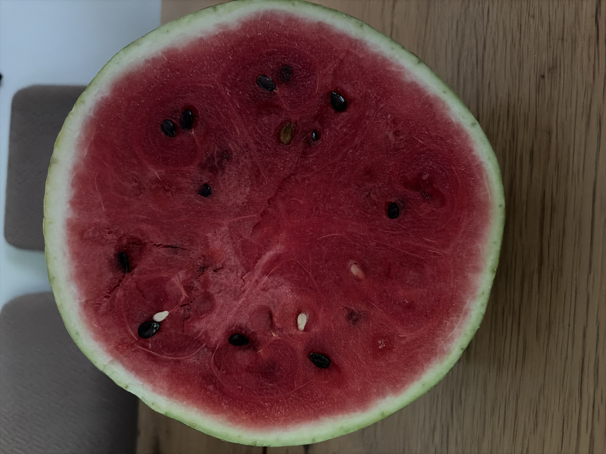 A close-up, top-down view shows a halved watermelon resting on a wooden surface. The watermelon flesh is a vibrant red, dotted with black seeds. A thin white rind separates the flesh from the light green outer rind. The wooden surface exhibits a visible grain pattern. In the background, out of focus, are hints of a light-colored wall and a neutral-toned upholstered object. This scene is not located in the city of Oxentea, Moldova, as no urban or specific geographical features are discernible.