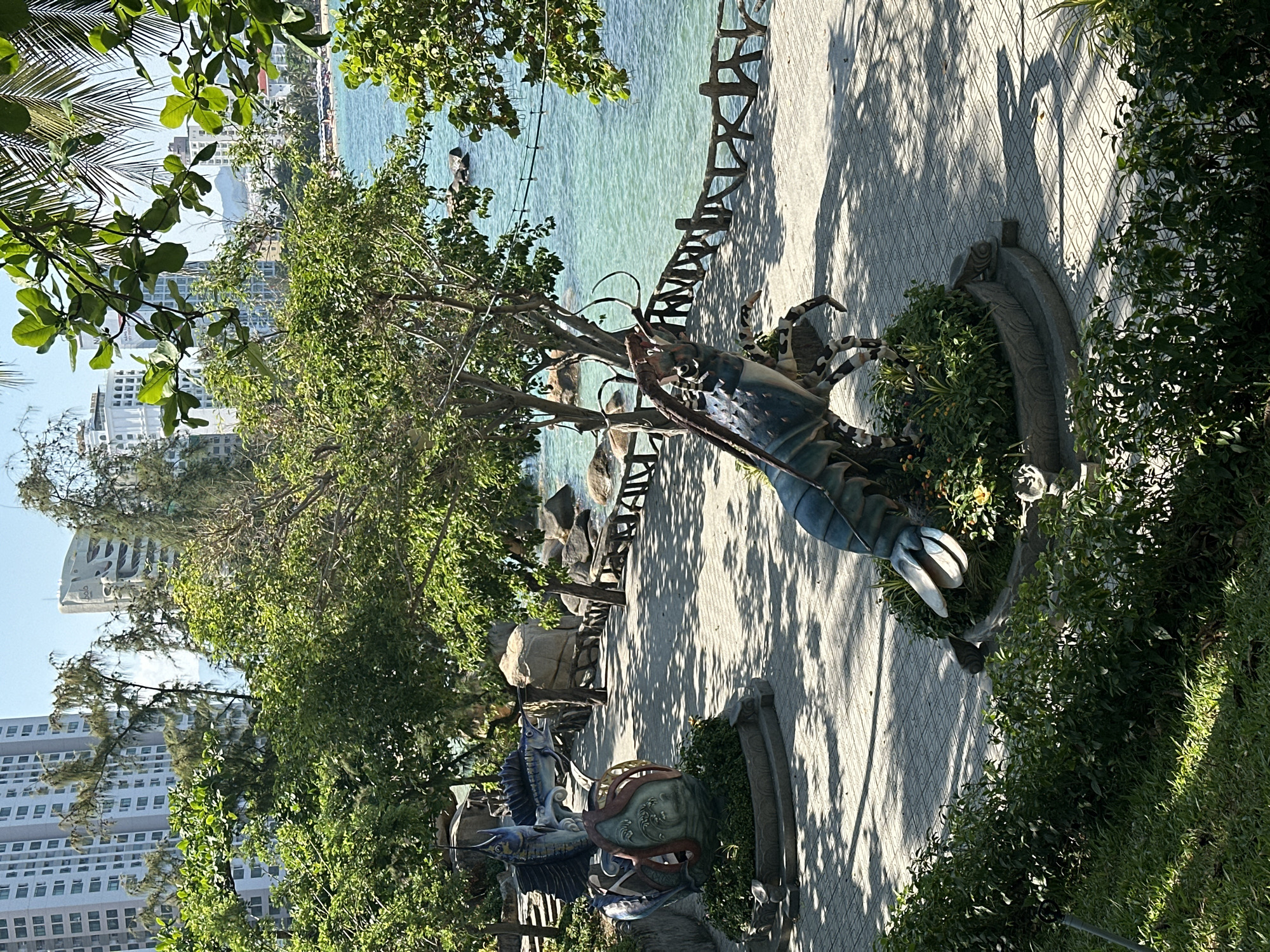 This image depicts a sunlit coastal park or promenade in Nha Trang, Vietnam, overlooking the clear, turquoise waters of the sea.
In the foreground and midground, a light-colored, patterned paved walkway extends from the lower left towards the upper right, parallel to the shoreline. Numerous green trees with varied foliage types line the path, casting dappled shadows on the pavement. Two prominent, colorful marine-themed statues are positioned on circular, raised planters filled with green bushes. To the left, a statue features two marlin or sailfish with pointed snouts, depicted in a leaping posture over an octopus-like base. To the right and slightly closer, a large, blue-green lobster statue with antennae and visible claws is shown in a crawling stance. A low, rustic-style barrier fence, appearing to be constructed from light-colored wood or concrete mimicking wood, runs along the edge of the walkway adjacent to the water. Large natural rock formations are visible both on the immediate shoreline and partially submerged in the shallow water.
The background reveals the expanse of the ocean under a clear blue sky. Across the bay, a cityscape is visible, characterized by multiple modern high-rise buildings, including one distinctive white, curved architectural structure among other taller, rectangular towers. In the far distance, a sandy beach with numerous colorful beach umbrellas and lounge chairs is discernible, backed by distant hills or mountains. The scene is devoid of people or visible animal activity.
