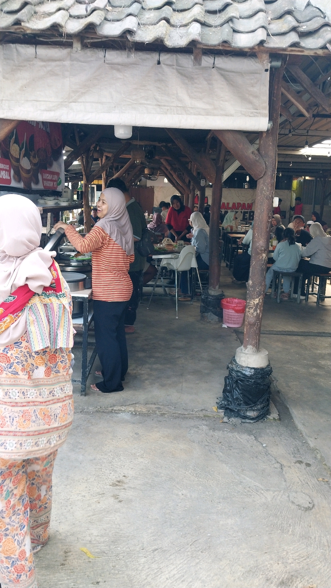 The image depicts an outdoor dining area at what appears to be a restaurant or warung in Pasuruan, Indonesia. The scene is an open-air structure with a tiled roof supported by wooden beams. In the foreground, two women are visible. The woman on the left is wearing a pink hijab and a floral-patterned outfit. The woman on the right, also wearing a hijab, is in an orange and white striped shirt and dark pants, and is interacting with a food preparation area. Several other people are seated at tables in the background, engaged in eating and socializing. The environment is rustic, with concrete flooring and visible building materials. There are tables, chairs, food displays, and food service items present. A red trash bin is also visible. A sign with Indonesian text is partially visible on the left side.