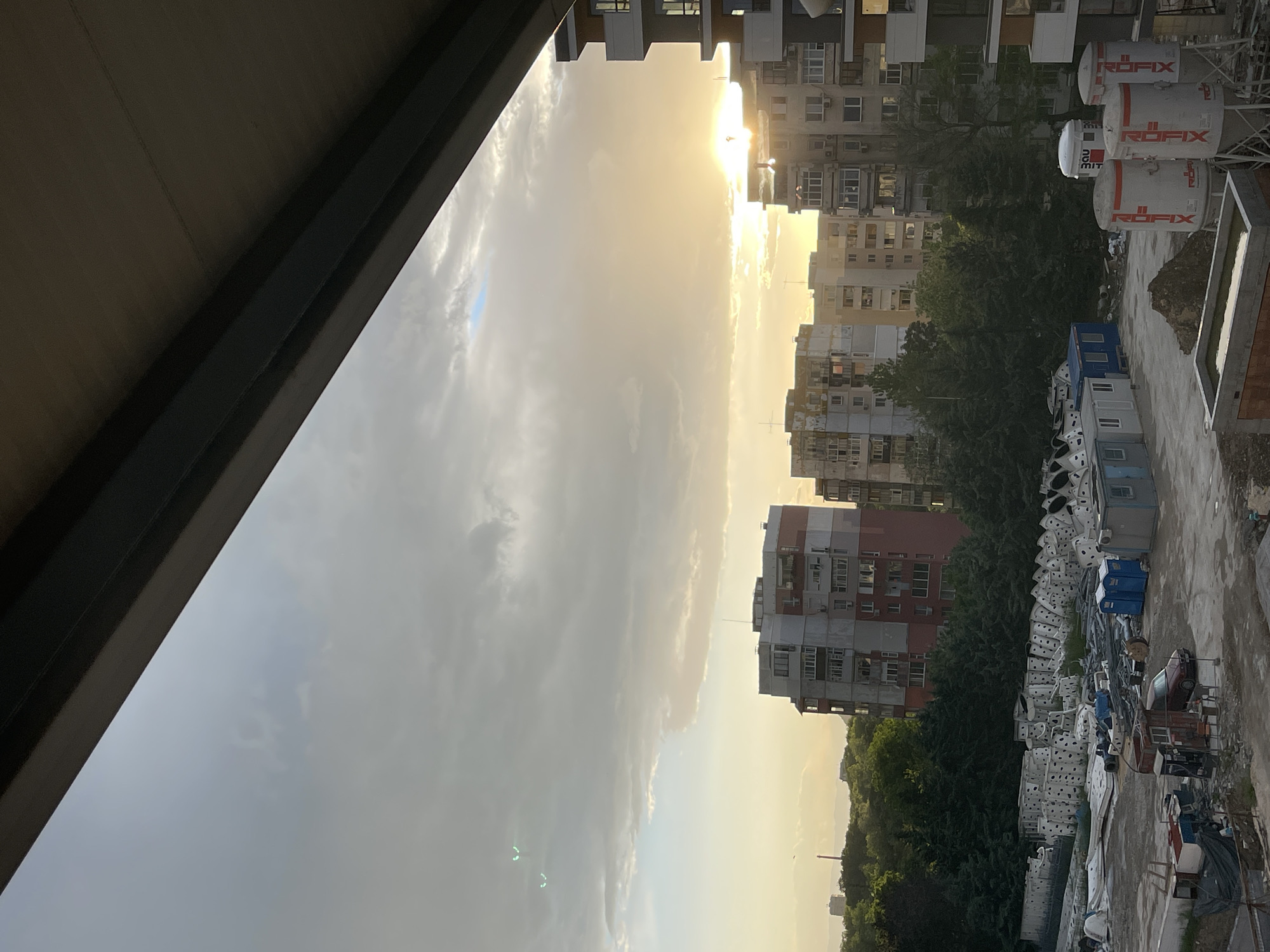 The image depicts an urban scene, likely taken from a high vantage point, possibly a balcony. The sky is partially obscured by clouds, with the sun visible behind them, casting a warm glow. Several multi-story residential buildings are visible, typical of urban construction in Plovdiv, Bulgaria. A construction site or storage area is located closer to the viewpoint, featuring containers, construction materials, and various objects covered in white dotted tarps. The presence of trees and other greenery suggests this location is in an area with some vegetation. The architecture and general layout are consistent with the urban landscape of Plovdiv.