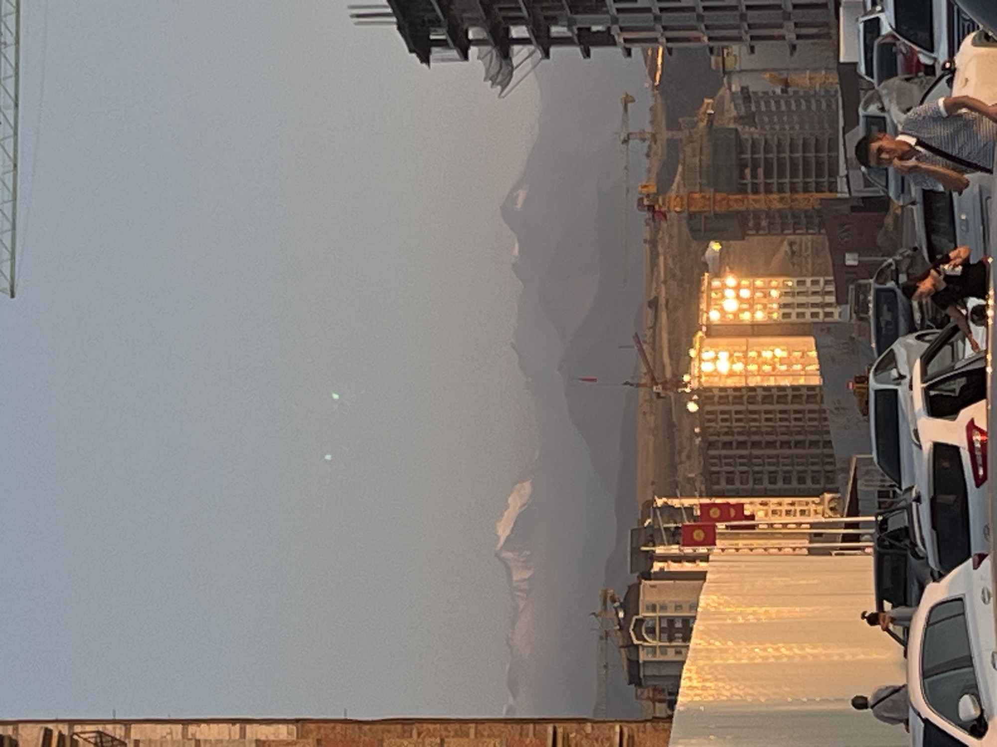 In the city of Bishkek, Kyrgyzstan, this image captures a city skyline at dusk, featuring several multi-story buildings under construction, some with lights illuminating their windows. A crane is visible above a building on the right. In the foreground, several cars are parked or in motion, with some people standing near them. A white wall with a textured surface is on the left side of the image. In the background, a mountain range with snow-capped peaks is visible under a hazy sky. Two small, blurry green lights are present in the upper part of the sky.