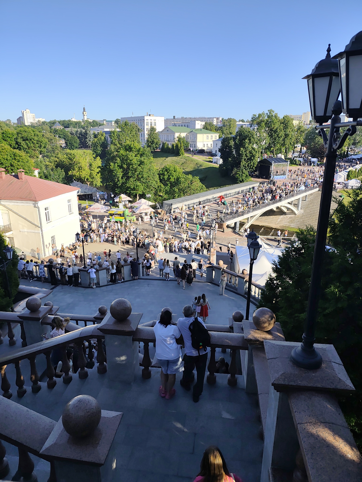 The image depicts a bustling outdoor event on a clear, sunny day, likely late afternoon or early evening given the warm lighting and long shadows. The scene is set in Vitsebsk, Belarus, and features a large gathering of people on a bridge and in an open area below.
In the foreground, a grand stone staircase with ornate railings and spherical finials leads down towards the activity. Several people are seen on the stairs, observing the event. Behind the staircase, a large crowd congregates in a plaza area, with many tents and stalls set up, suggesting a market or festival. A prominent feature is a long, arched bridge that spans over a lower area, filled with people walking across it. Further in the background, several buildings, including some multi-story white structures and a church tower, are visible amidst lush green trees, indicating an urban park or city center setting.
The overall atmosphere appears lively and festive, with many individuals casually dressed and mingling. The warm sunlight casts a pleasant glow, contributing to a cheerful mood. The presence of a stage and tents suggests a public gathering, possibly a cultural festival or a local celebration. No specific text is clearly legible in the image, though some signs are visible on the tents and on the green food truck.