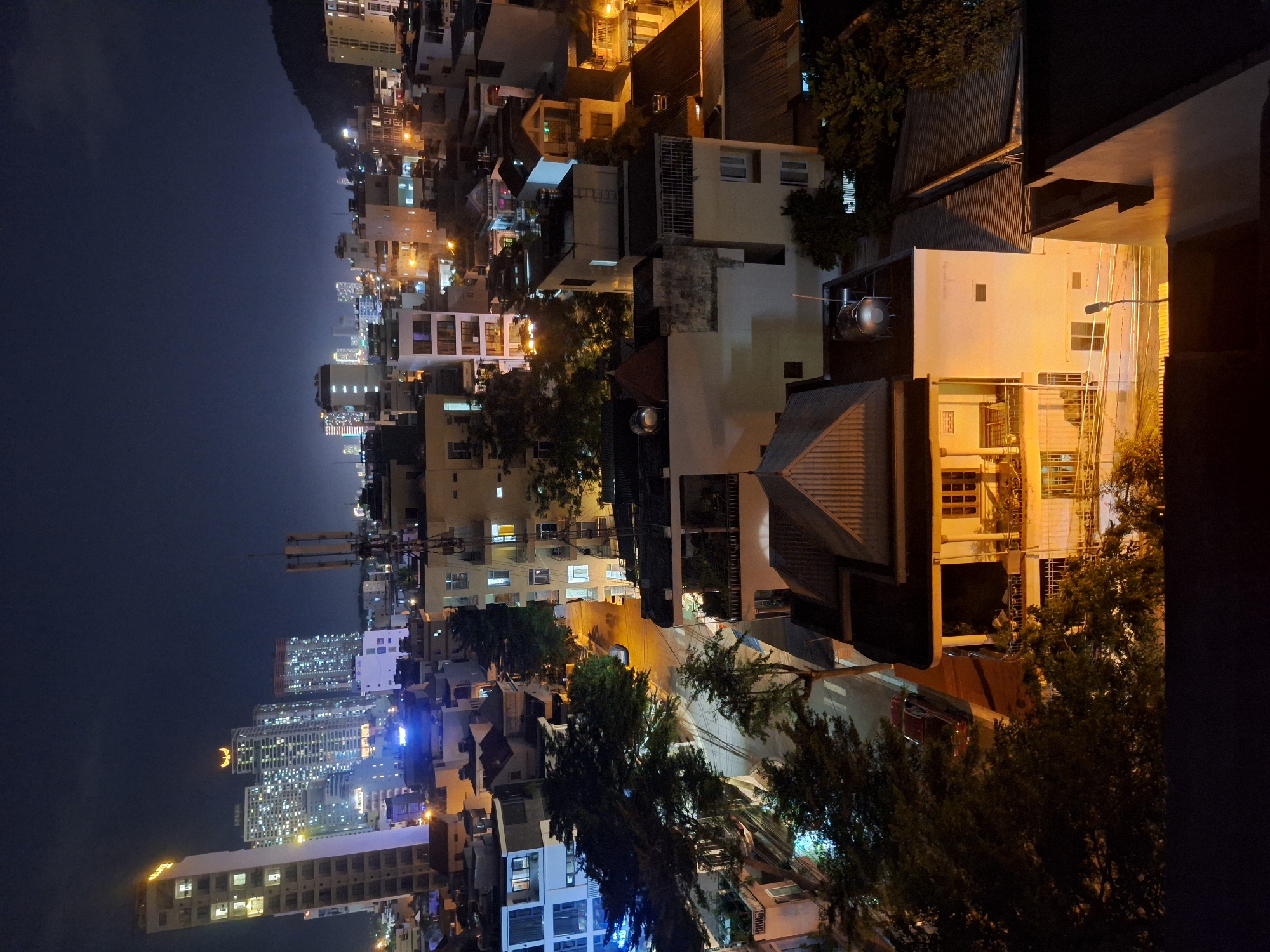 This is a nighttime aerial photograph taken in Nha Trang, Vietnam, looking down on a densely populated urban area with a mix of residential and commercial buildings. The scene is illuminated by artificial lights from buildings, streetlights, and some visible signage. Tall, modern apartment buildings with illuminated windows dominate the background, interspersed with shorter, older structures. A winding street runs through the middle ground, with trees lining its sides. A red car is parked on the street, and other vehicles are not clearly discernible. The foreground shows the rooftops of several buildings, including one with a prominent triangular roof and another with a water tank. The overall impression is of a bustling city after dark, with the lights creating a warm, ambient glow against the dark sky. The presence of multiple apartment complexes suggests a residential district within the city. The distant hills in the background indicate the city's coastal location.