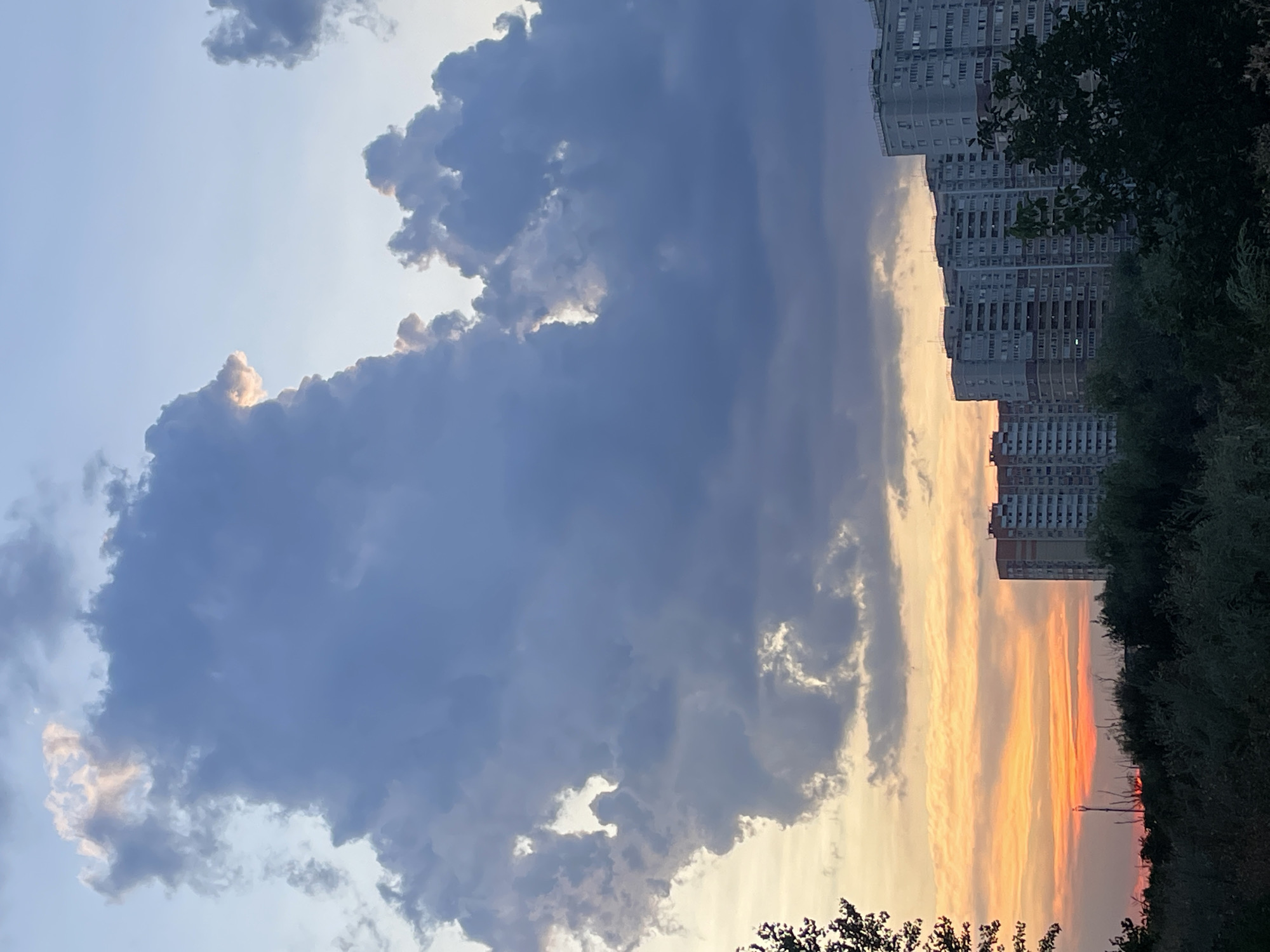 The image depicts a cityscape at sunset. Dominating the upper portion of the frame are large, dark gray clouds, with a sliver of light peeking through the top center, suggesting a break in the clouds or the sun's rays. In the lower half of the image, a row of tall, multi-story apartment buildings is visible, characterized by their light gray facades and numerous balconies. These buildings are set against a backdrop of a warm-toned sky, transitioning from orange and yellow near the horizon to a pale blue above. In the foreground, a dense band of dark green foliage, likely trees and bushes, obscures the lower parts of the buildings and the ground. The overall scene is one of urban twilight, with the natural elements of the sky and vegetation contrasting with the man-made structures. The image does not contain any people or discernible actions.