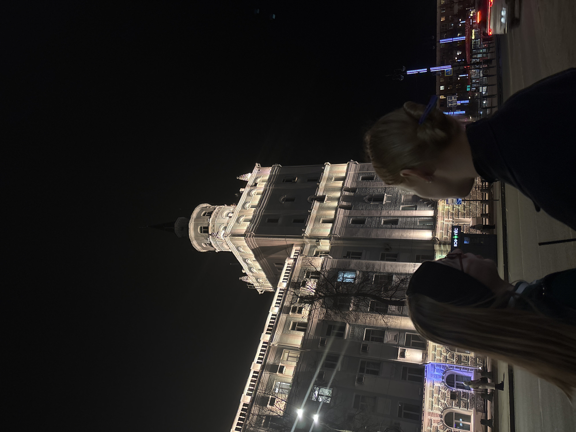 At night in Perm, Russia, two individuals are positioned on the sidewalk, viewing a large building. The building is lit, featuring a tower and a series of windows and architectural details. Street lights are visible. Traffic moves on the road in front of the building.