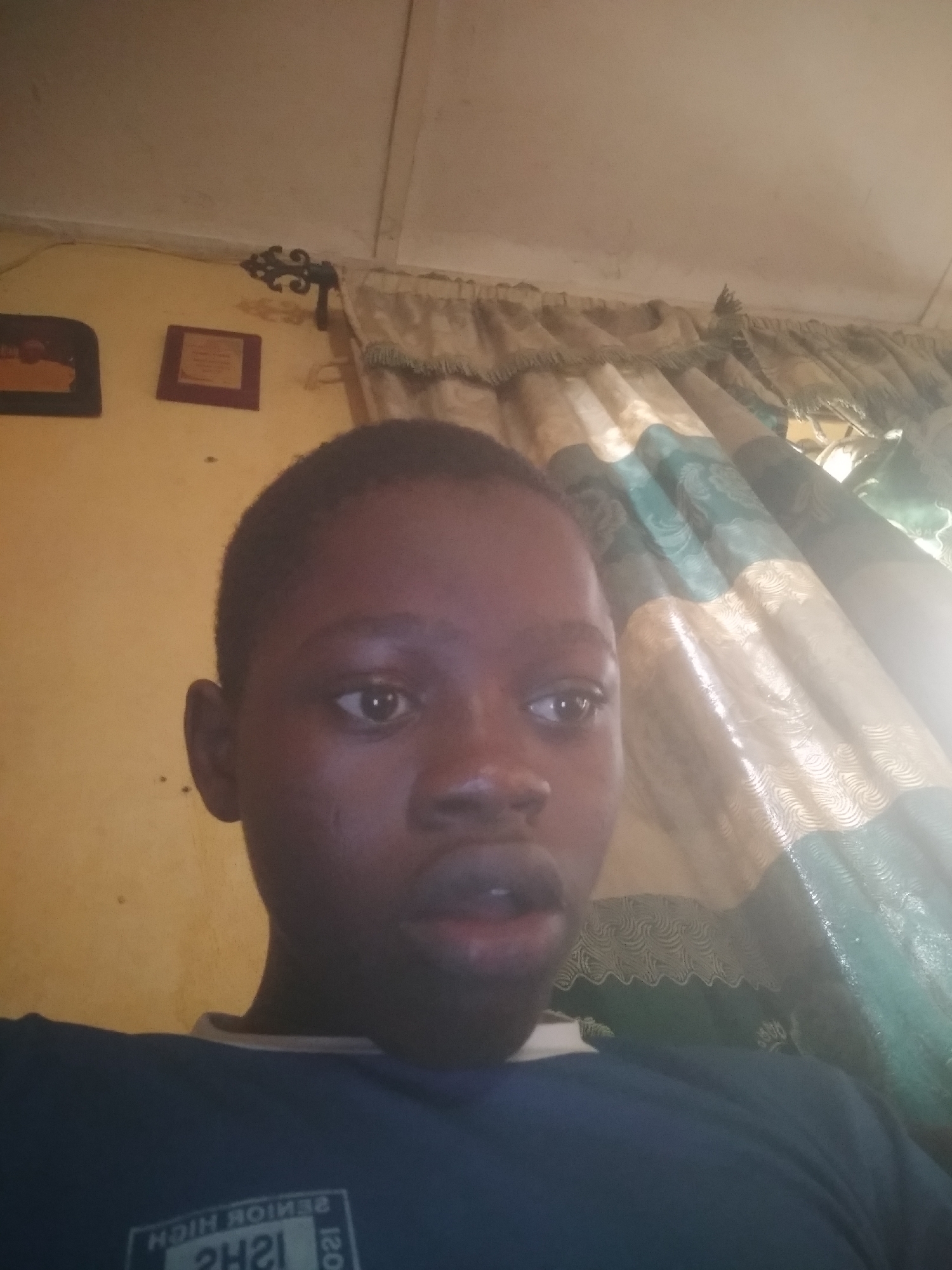 A head-and-shoulders shot shows a young person with dark skin and short, dark hair looking slightly to the right with a surprised expression. Behind the subject are wall decorations and curtains. The location is in the city of Unknown, Unknown.