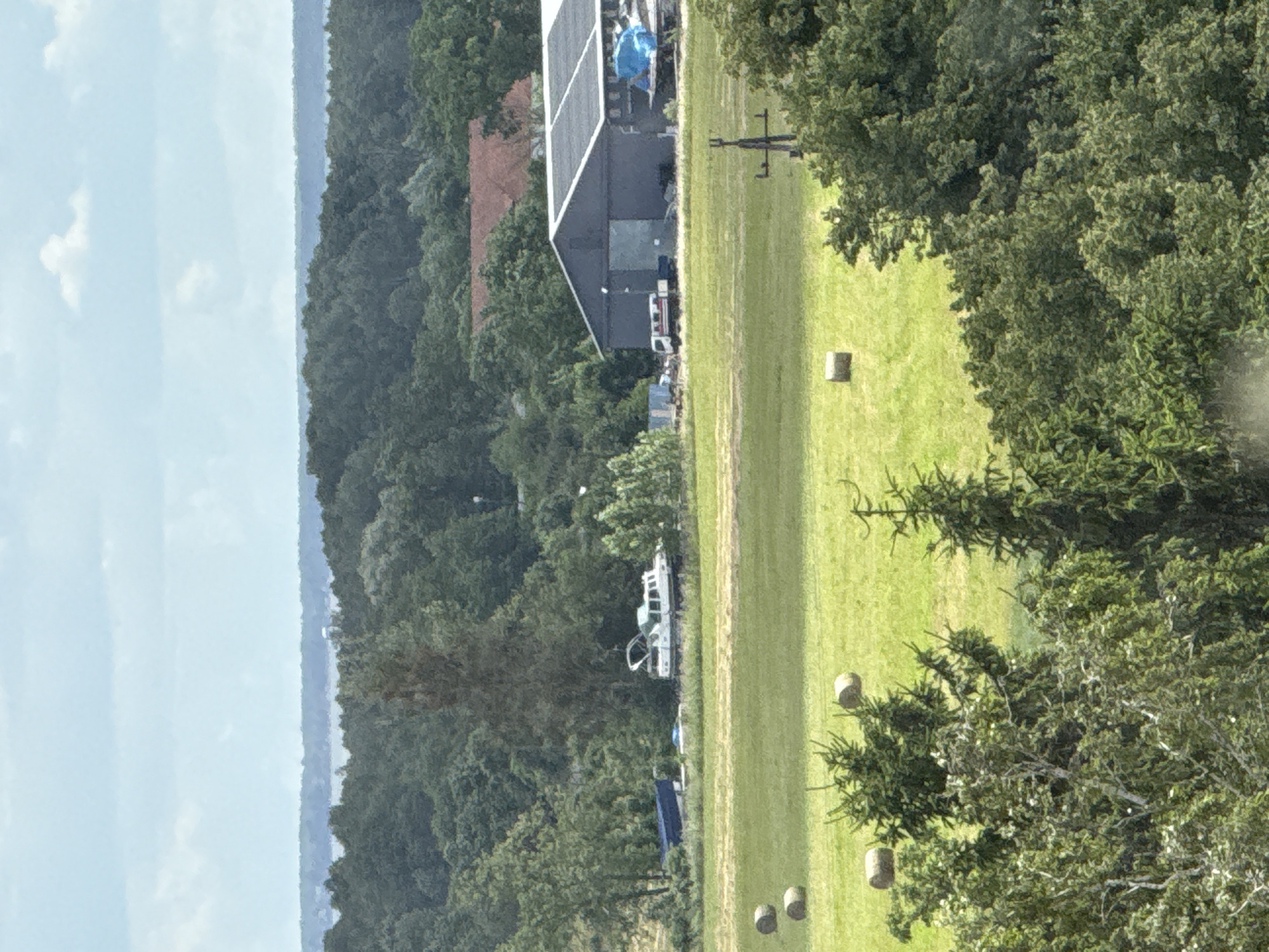 This image depicts a rural landscape with a body of water visible in the distance, likely part of Lake Mikołajskie in Mikołajki, Poland. In the foreground, a vibrant green field contains several round hay bales. Beyond the field, a large, dark-colored barn-like structure is visible, with an airplane and a boat partially covered in blue tarpaulin parked in front of it. A white truck with red and white stripes is also positioned near the barn. To the left of the barn, a white boat with an open top is situated on the grass, with another blue boat or cover nearby. The background is characterized by a dense forest of green trees, with a hint of a reddish-brown roof of a building peeking through the foliage. The sky is a pale blue with scattered white clouds, suggesting a daytime scene with moderate weather conditions. No people are visible in the frame.